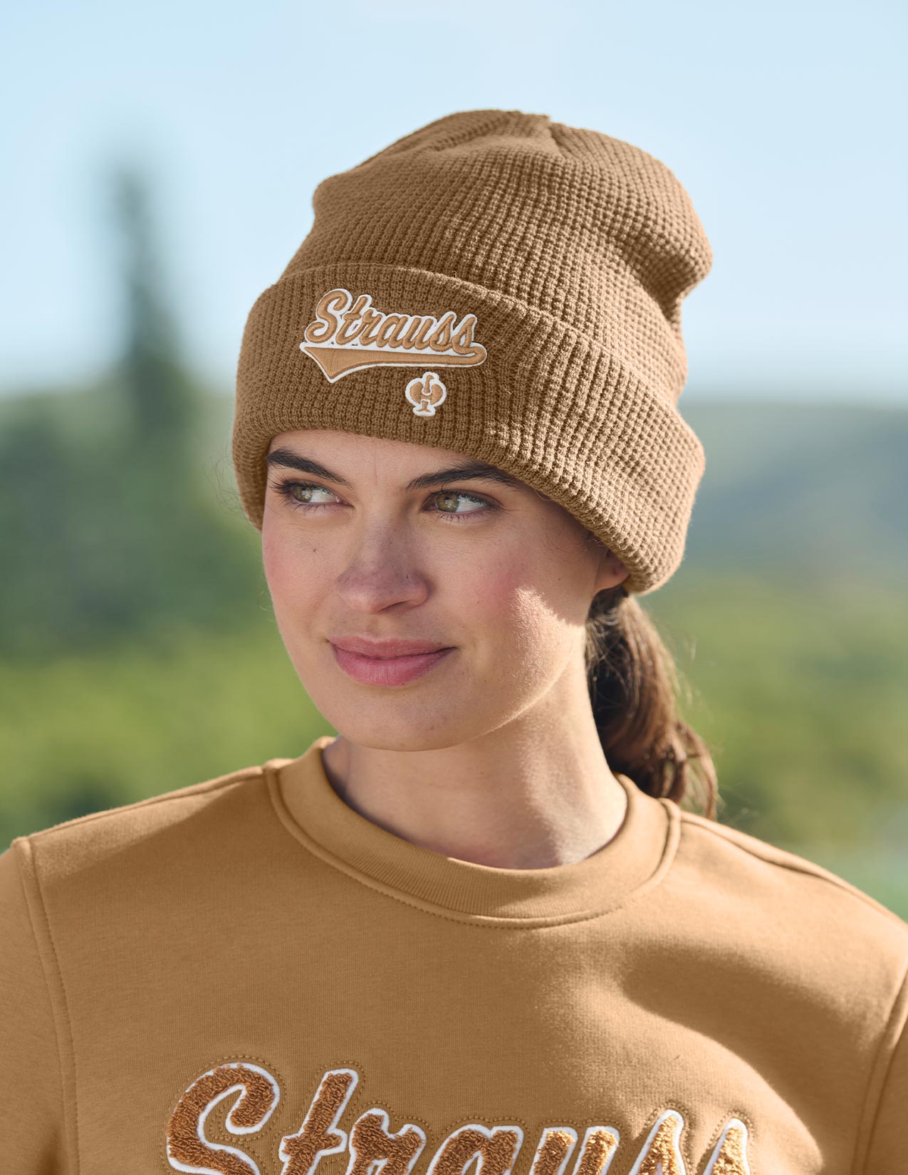 Woman wearing a brown knit beanie and matching sweatshirt with 'Strauss' text outdoors.