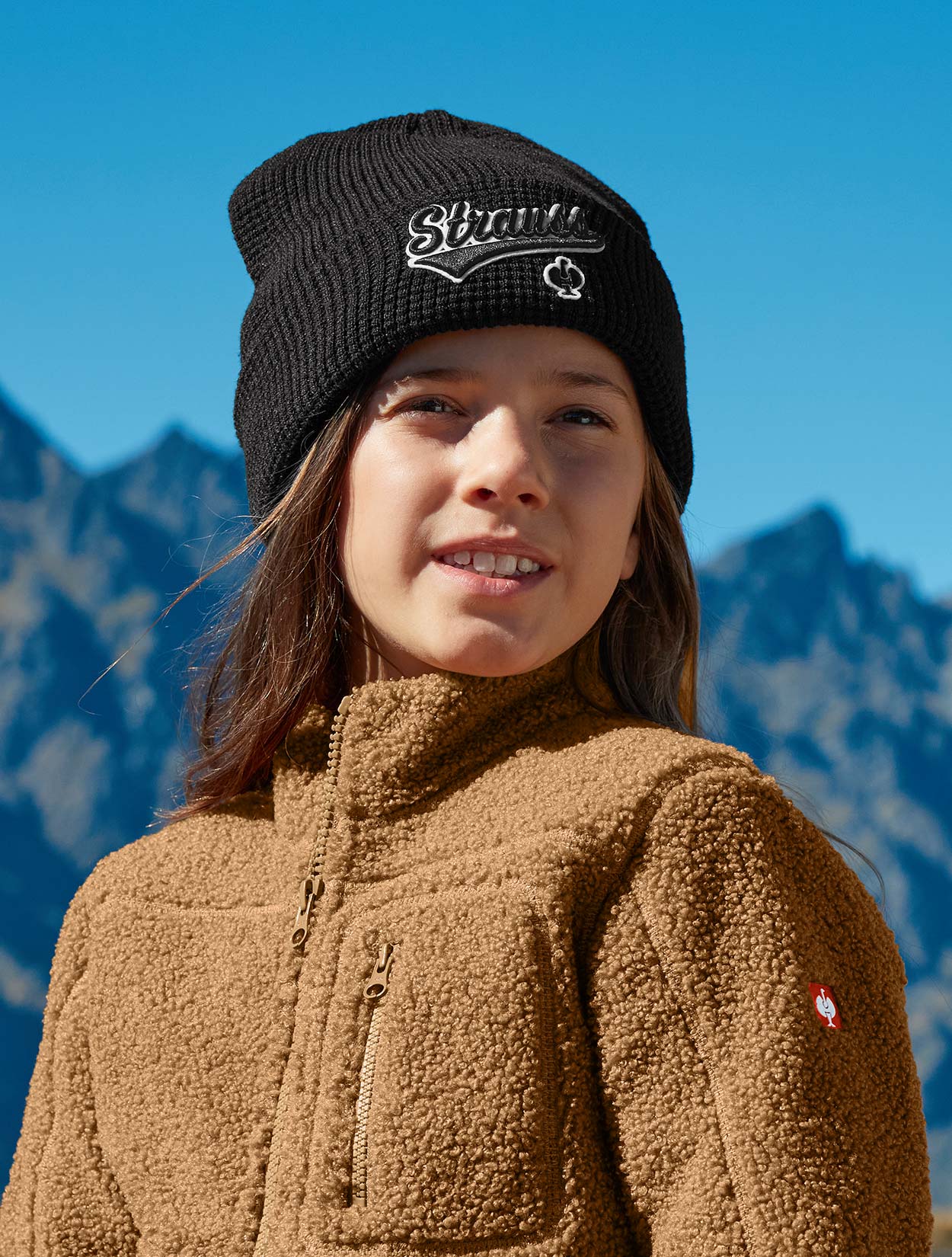 Child wearing a black knit beanie with a logo and a brown fleece jacket against a mountainous background.