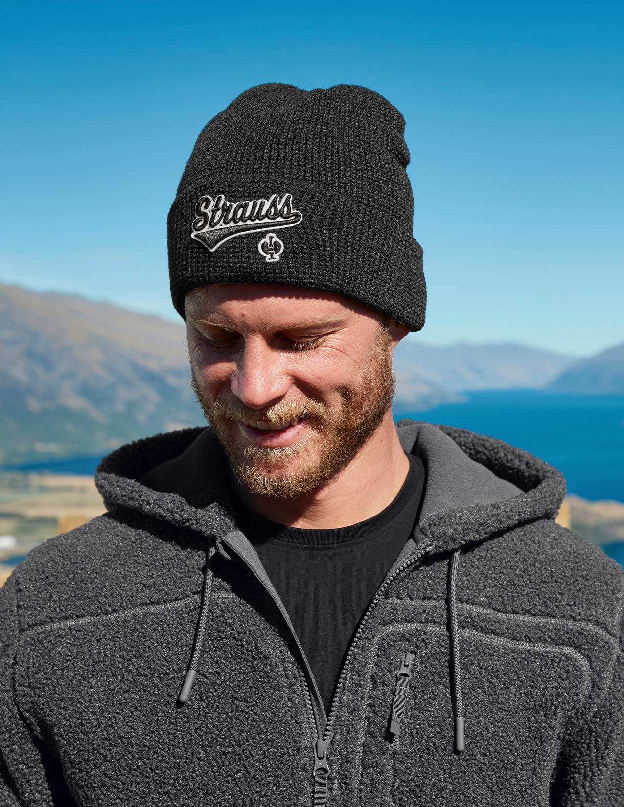 Man wearing a black beanie with 'Strauss' logo outdoors against a blue sky and mountains.