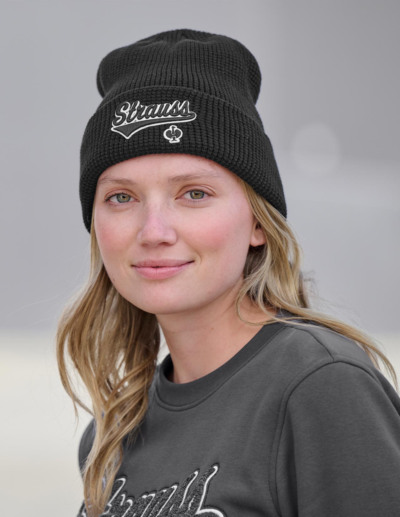 Woman wearing a black knit beanie with a logo and text, against a neutral background