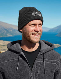 Man wearing a black knit beanie with a logo and gray fleece jacket outdoors with mountains and lake in the background.