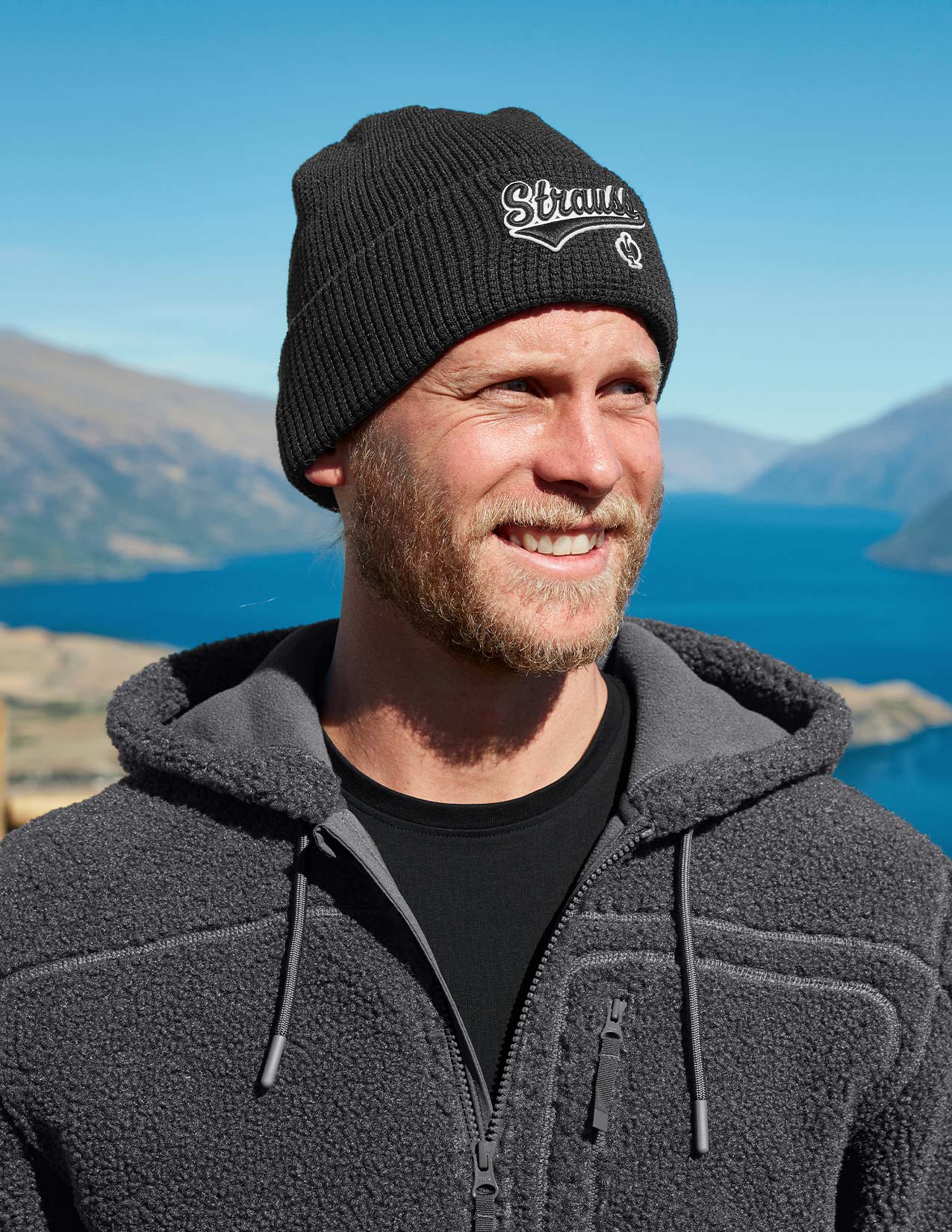 Man wearing a black knit beanie with a logo and gray fleece jacket outdoors with mountains and lake in the background.