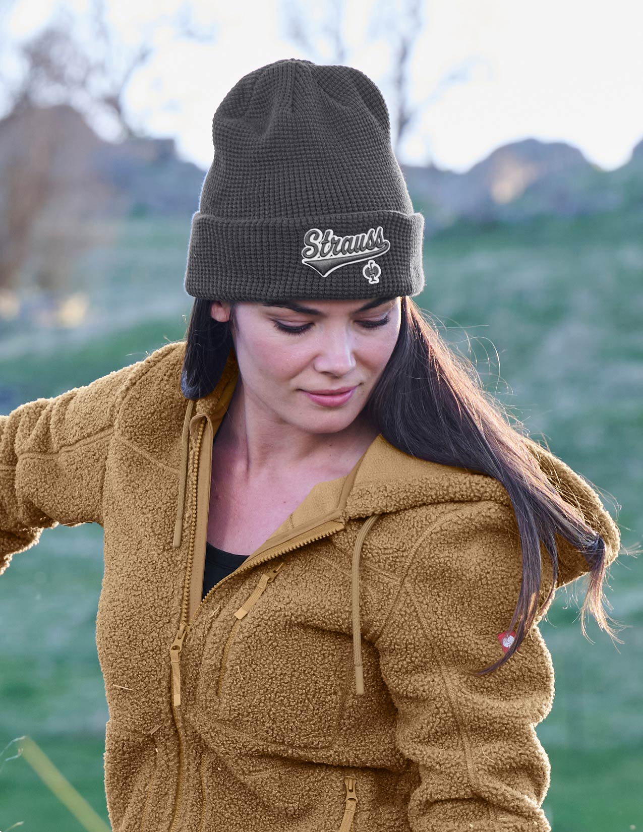 Woman wearing a brown jacket and grey beanie with 'Strauss' logo outdoors.
