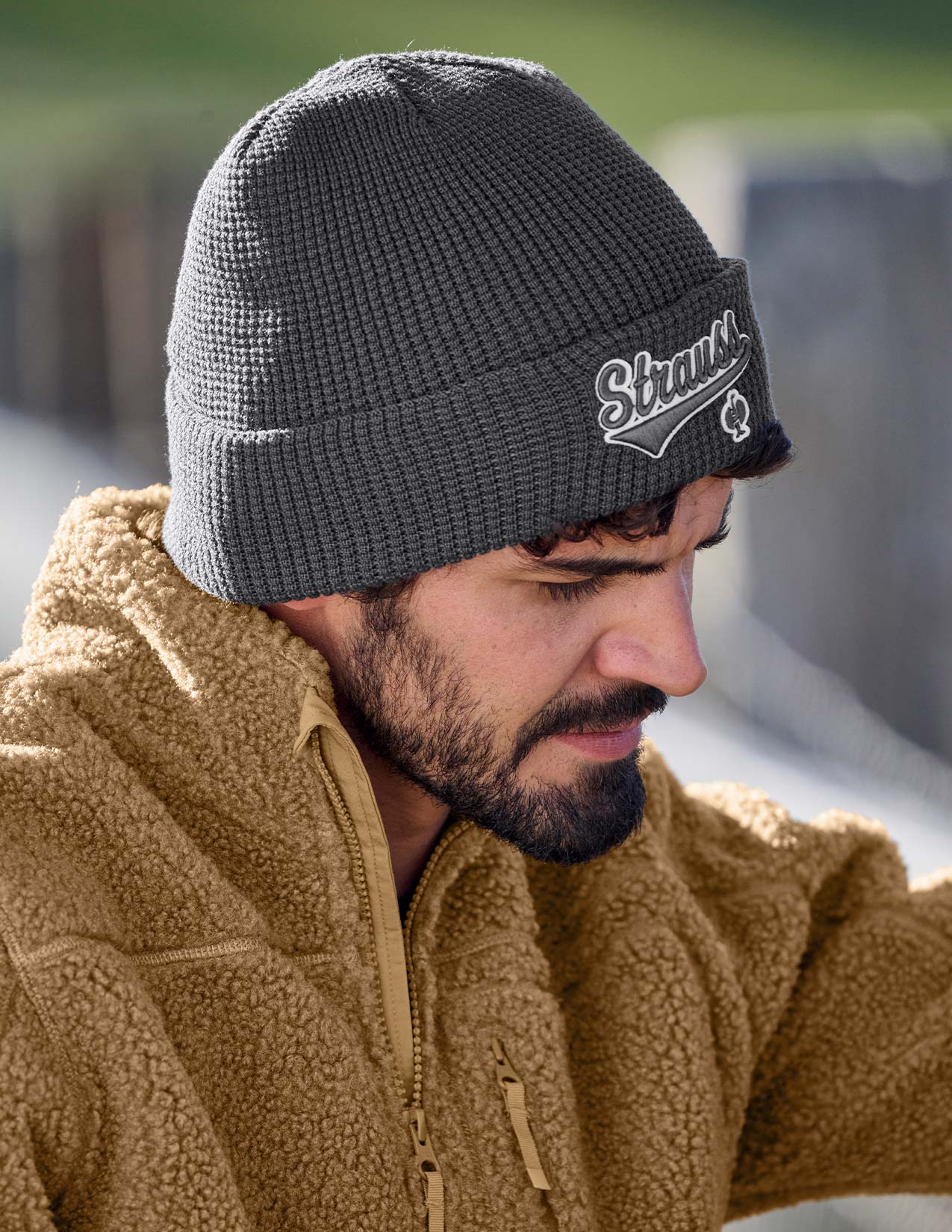 Man wearing a gray beanie with a logo and a brown jacket outdoors.