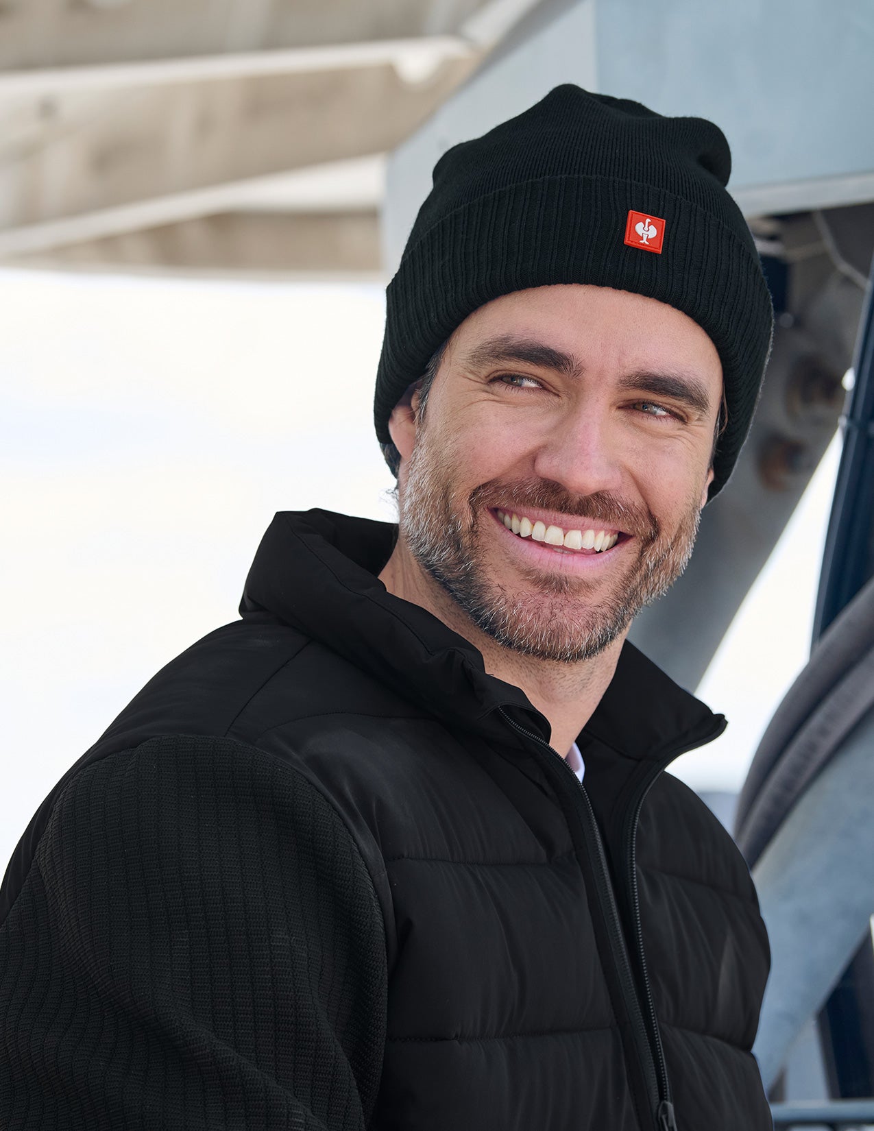 Man wearing a black beanie with a logo and black jacket, smiling outdoors.