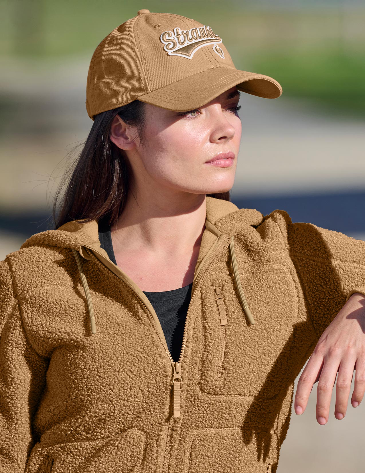 Woman wearing a brown cap and jacket with 'Strauss' branding, standing outdoors.