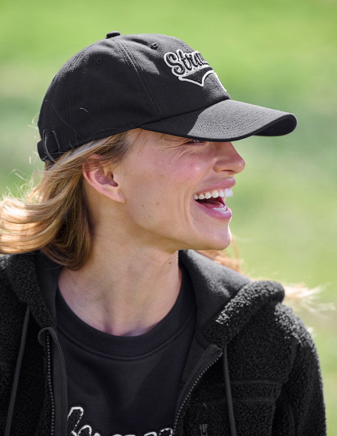 Woman wearing a black cap with 'Strauss' branding against a blurred green background