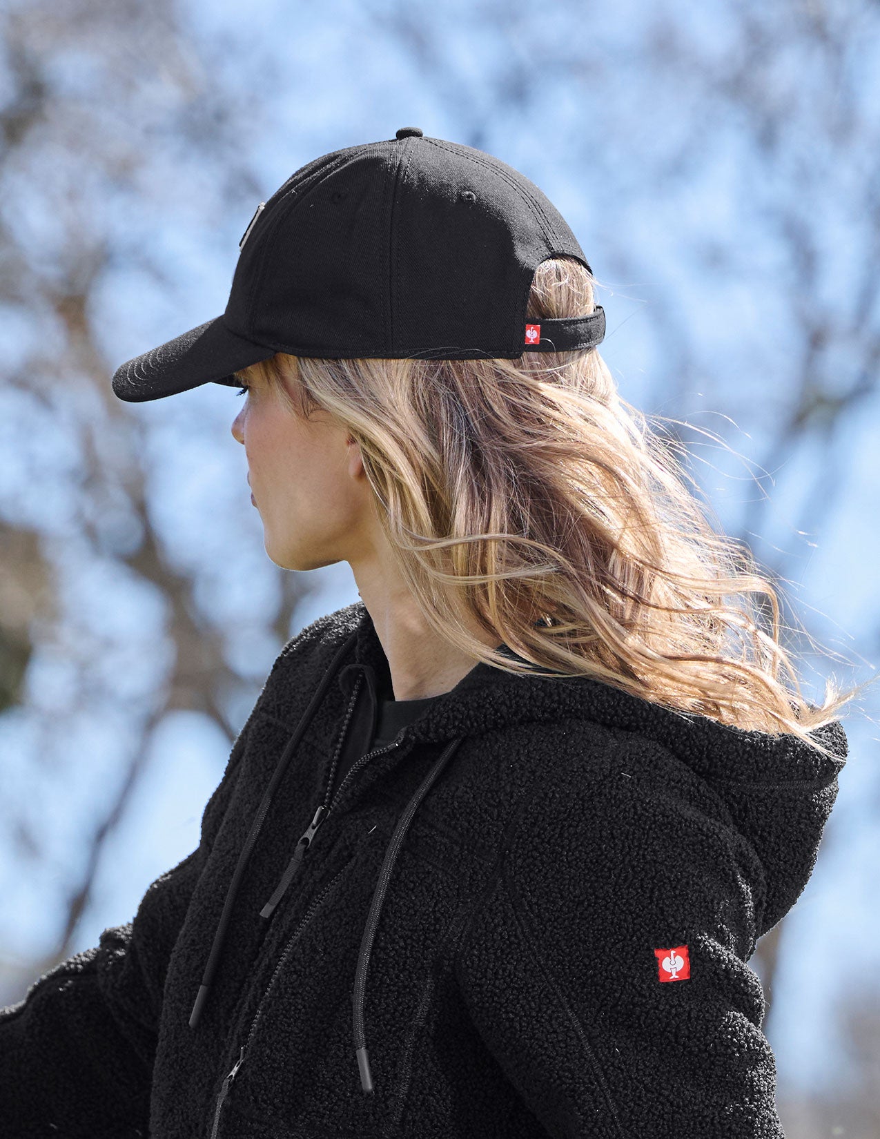 Woman wearing a black cap and black jacket with a logo, standing outdoors with blurred trees in the background.
