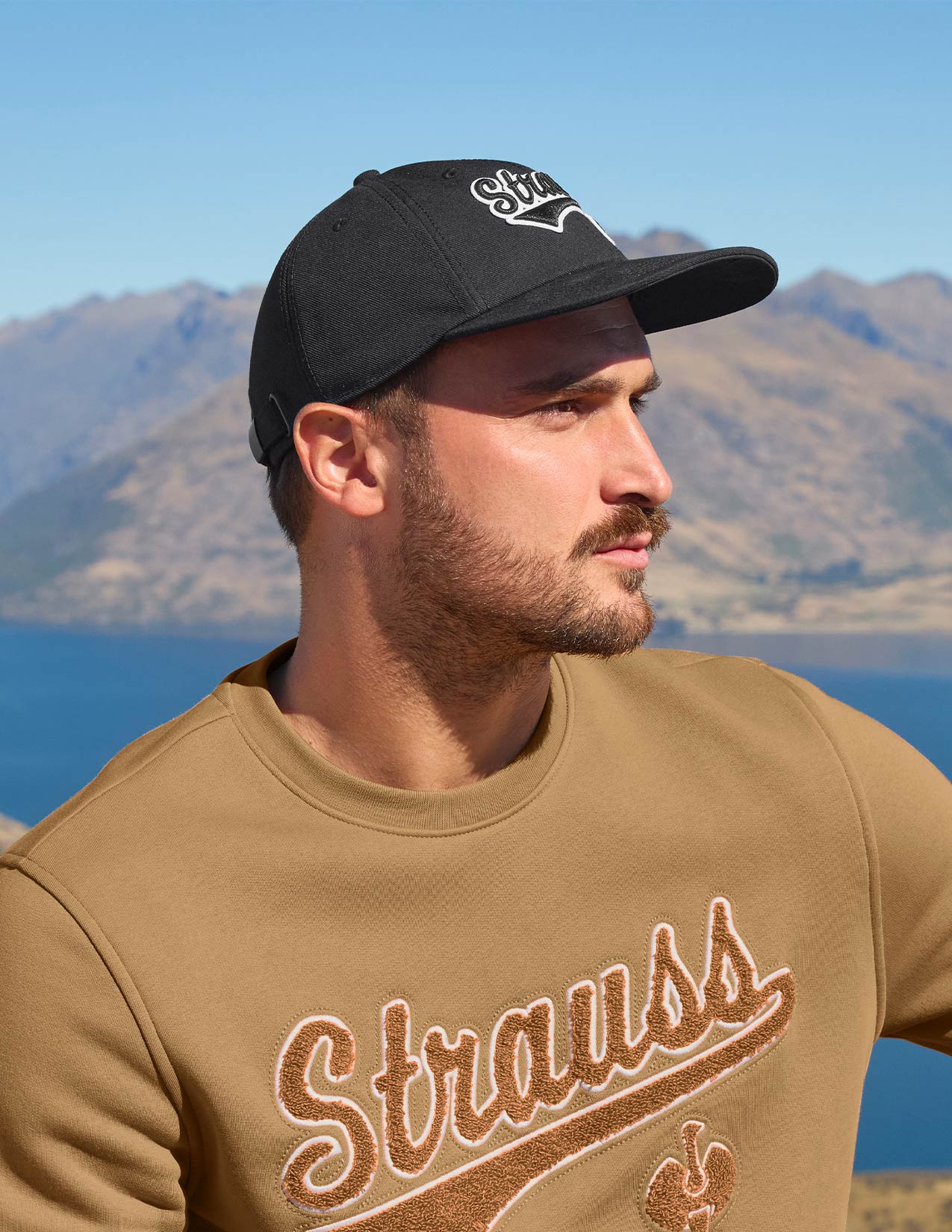 Man wearing a black cap and brown 'Strauss' sweatshirt with mountains and lake in the background