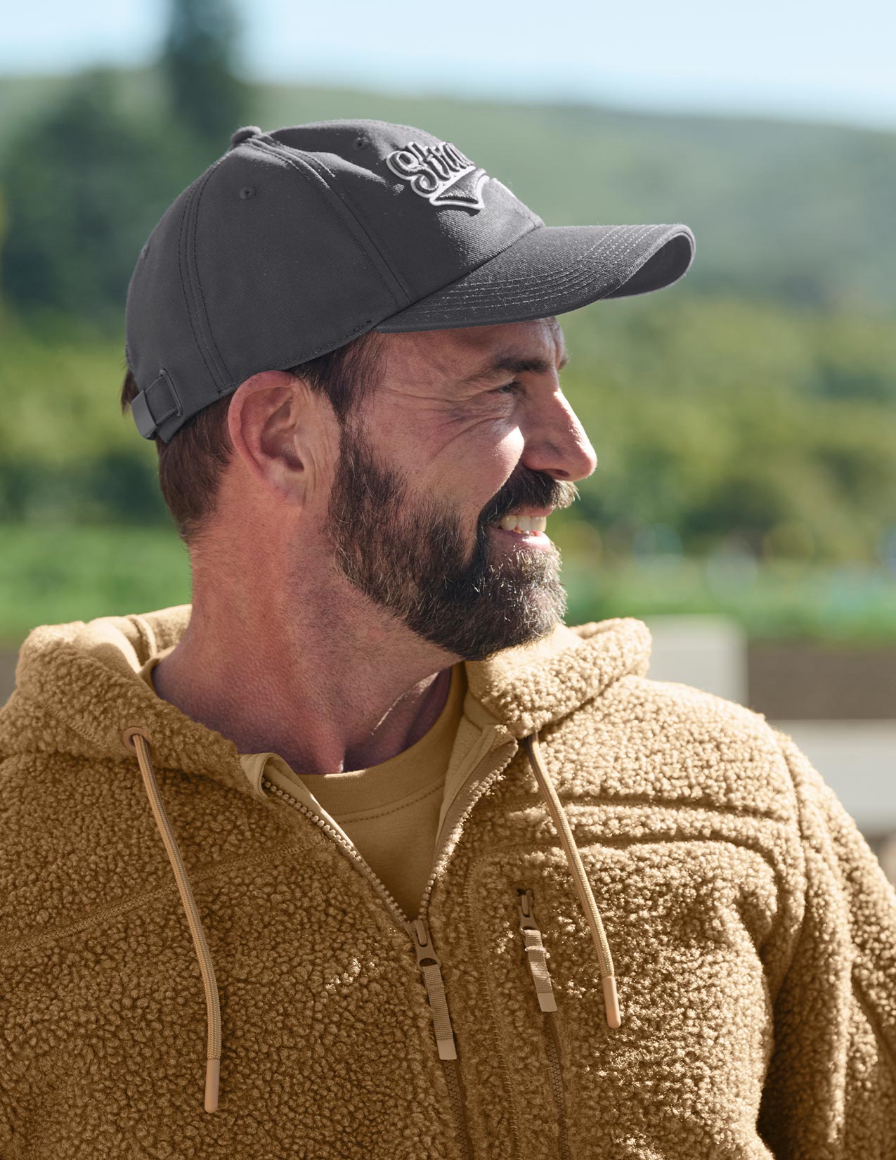 Man wearing a dark gray cap with a logo and a brown fleece jacket outdoors.