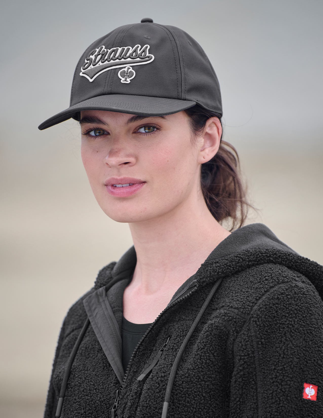 Woman wearing a dark gray cap with 'Strauss' logo on a neutral background