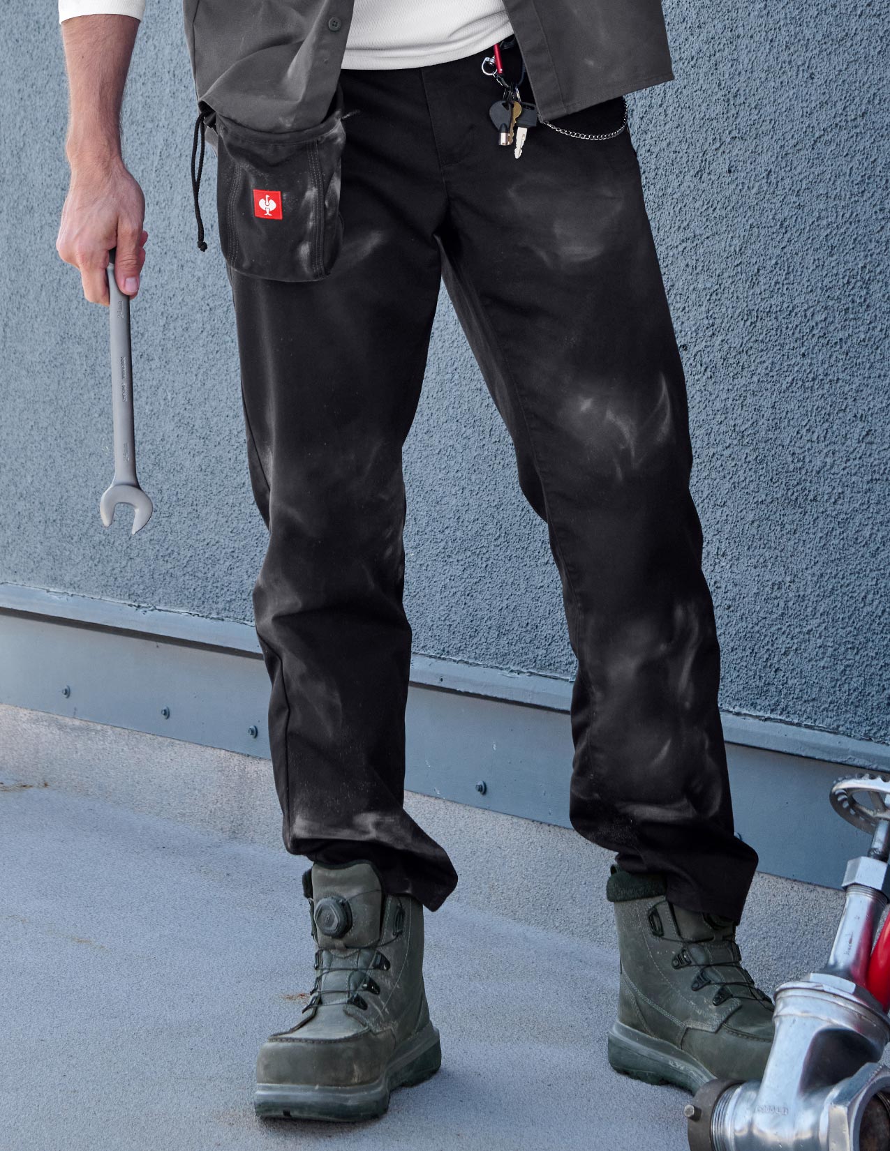 Person wearing black pants and boots holding a wrench against a gray wall.