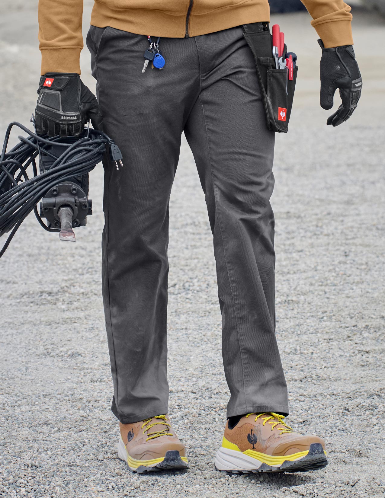 Person wearing gray work pants, brown shoes, and carrying tools on a gray pavement.