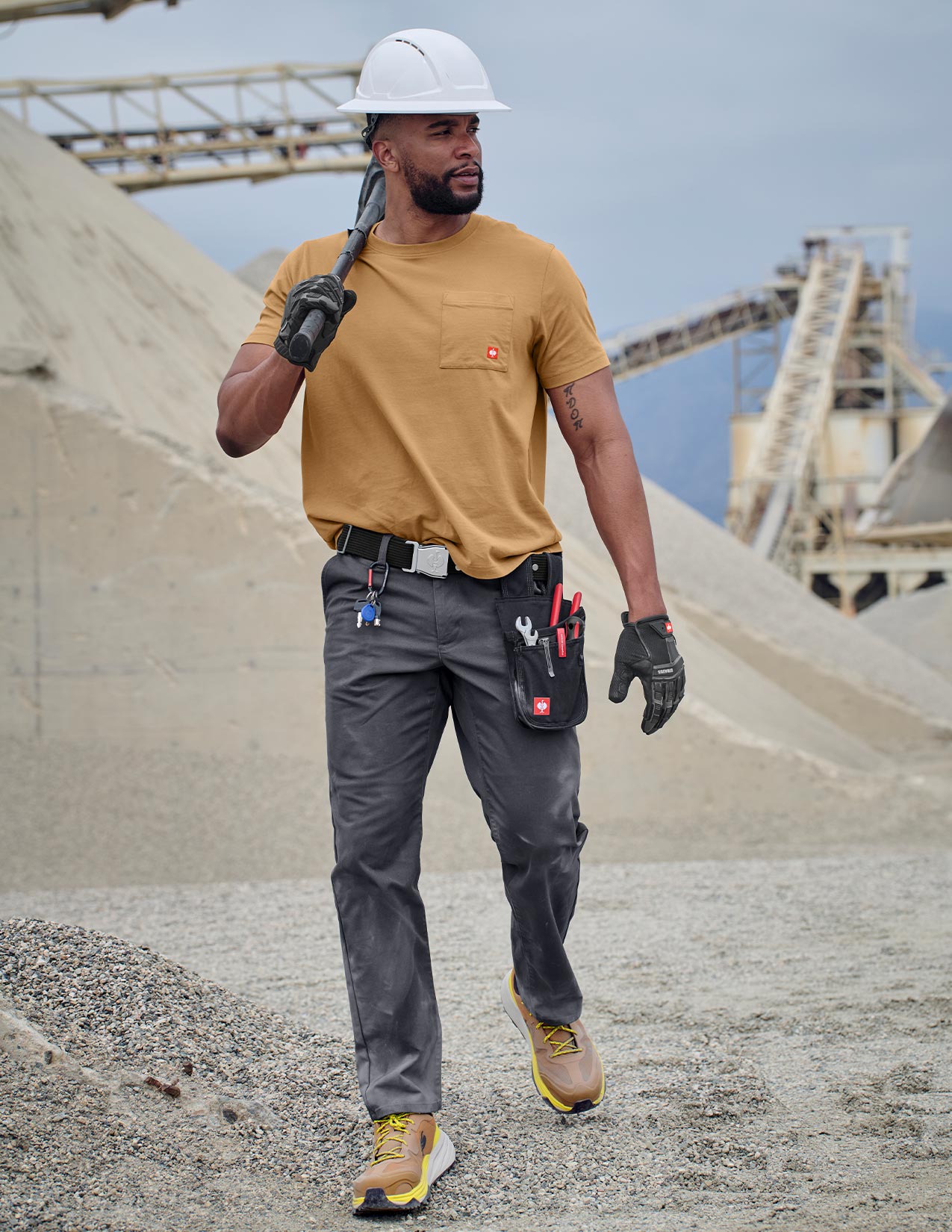 Man in construction attire with hard hat and tools walking on a construction site.