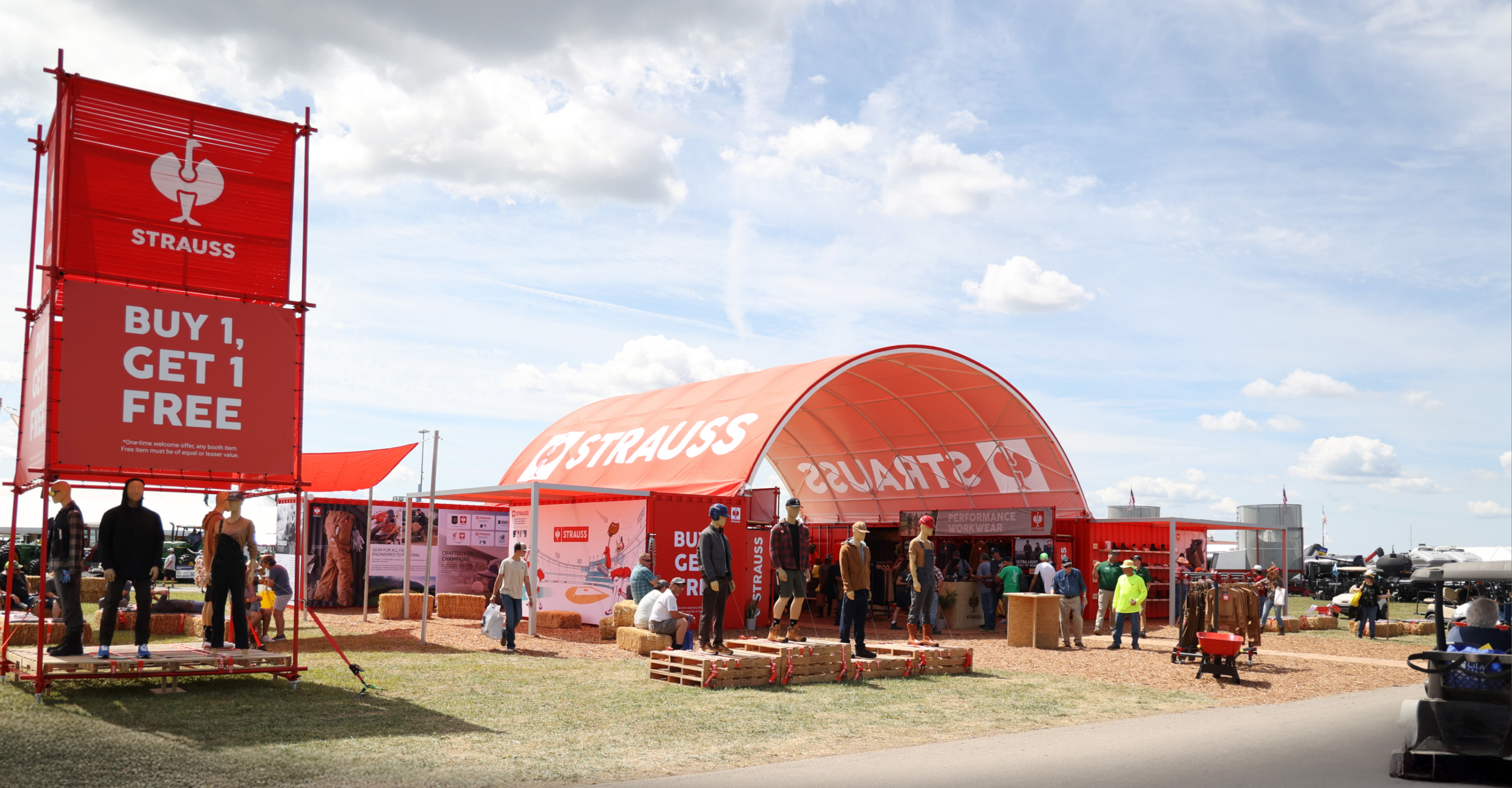 Outdoor Strauss booth with a red tent and BUY 1, GET 1 FREE sign. People are gathered around, some sitting on hay bales and pallets. The sky is partly cloudy and vehicles are visible nearby.