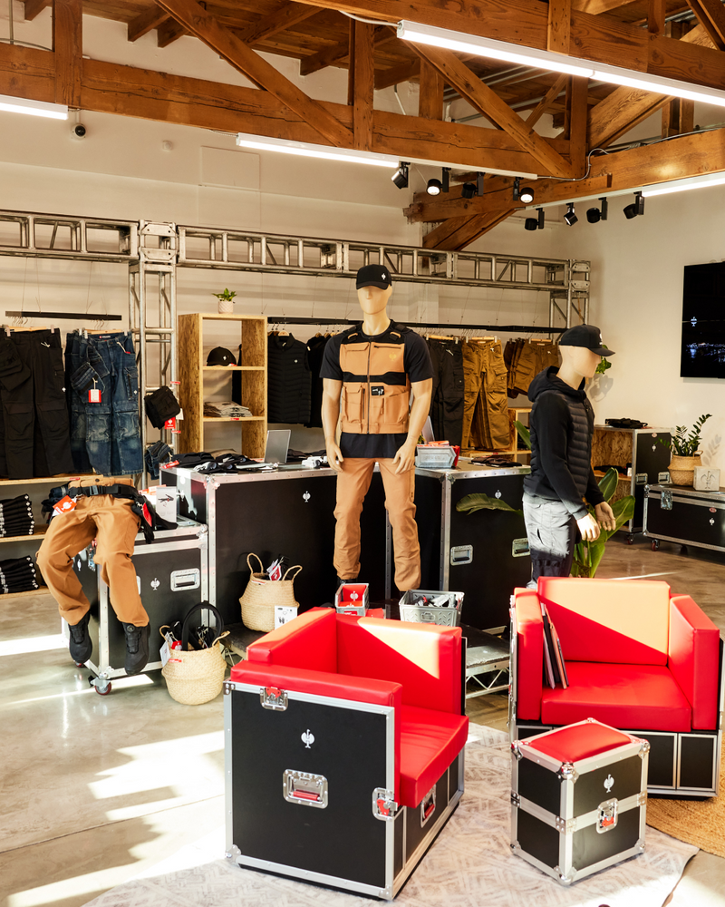 Clothing store interior with two mannequins wearing brown workwear and caps, surrounded by black and red chairs, display trunks, and clothing racks under exposed wooden beams and bright lighting.