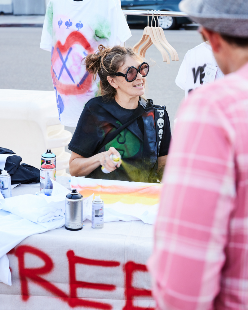 A woman with sunglasses spray paints a t-shirt at an outdoor booth. Art supplies and decorated shirts are on the table, and a person in a pink plaid shirt stands nearby.