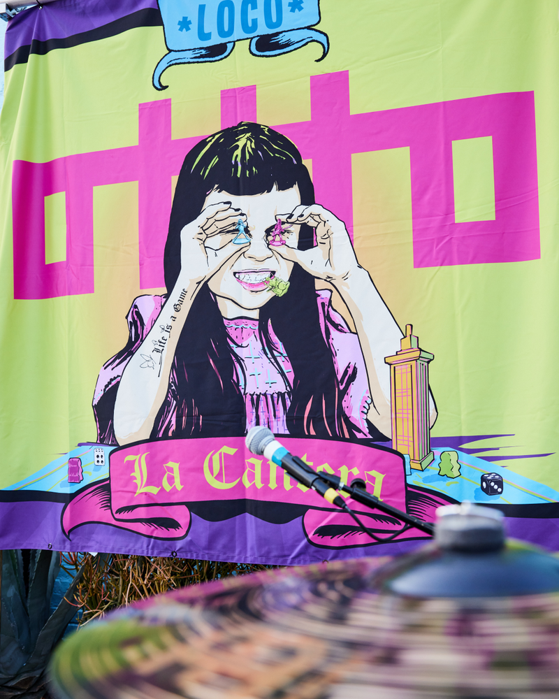 A colorful mural shows a girl holding candy over her eyes, with La Calera and Loco text, bold shapes, and vibrant colors in the background. A cymbal is blurred in the foreground.