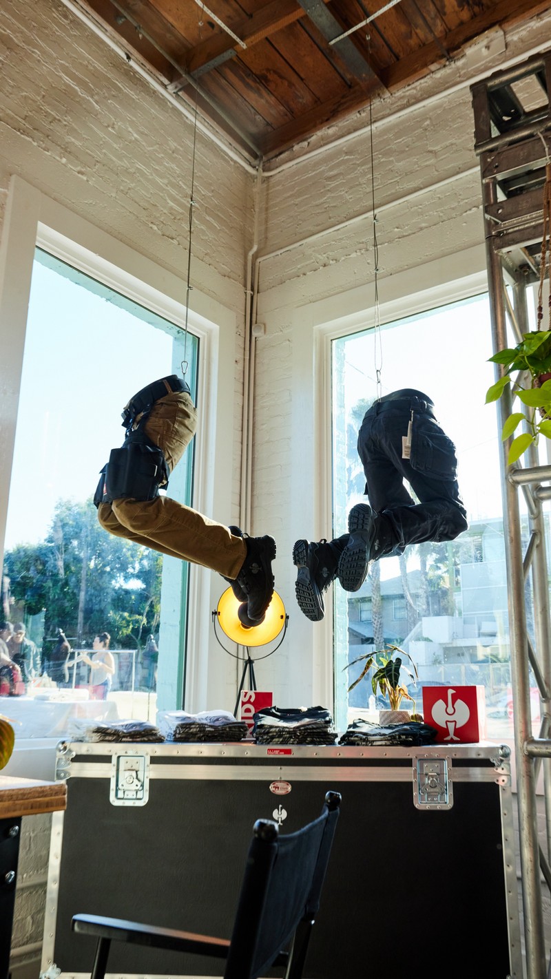 Two pairs of pants and boots, arranged as if seated and hanging in mid-air, are suspended by wires near large windows in a sunlit room with exposed wood ceiling beams.