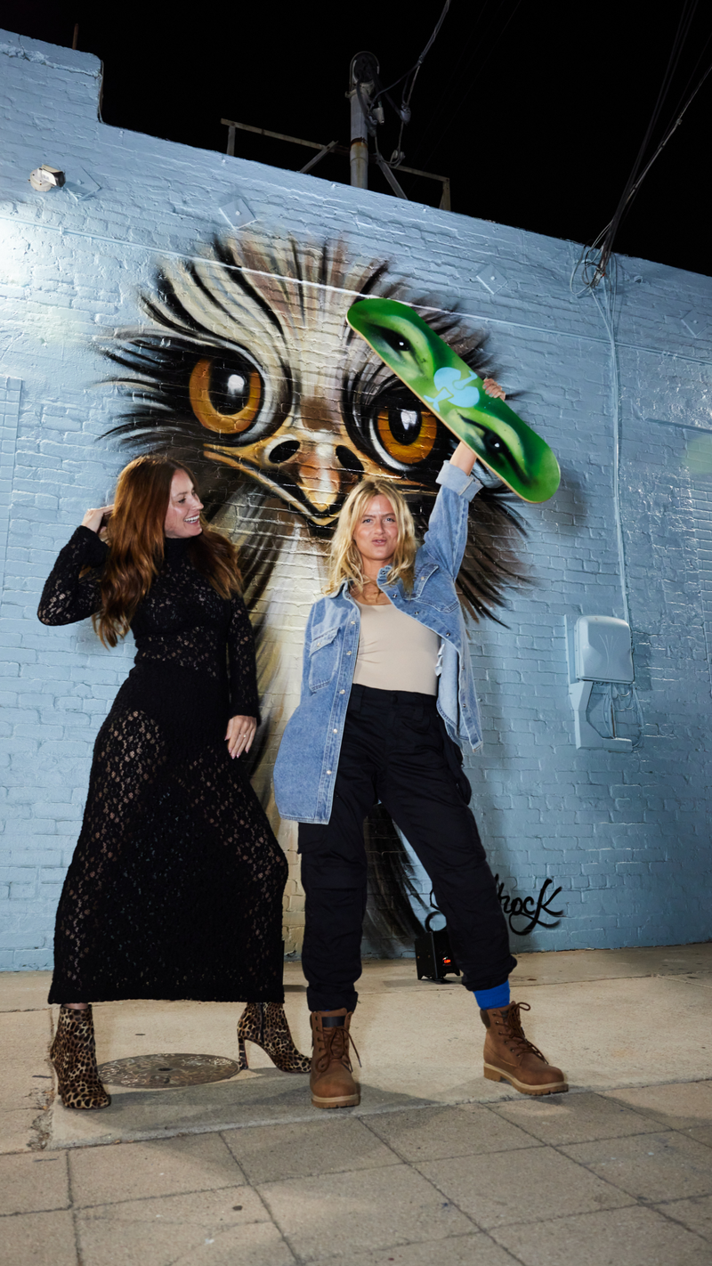 Two women pose in front of a mural of a wide-eyed owl; one wears a black dress, the other lifts a skateboard while wearing boots, dark pants, a beige top, and a denim jacket.