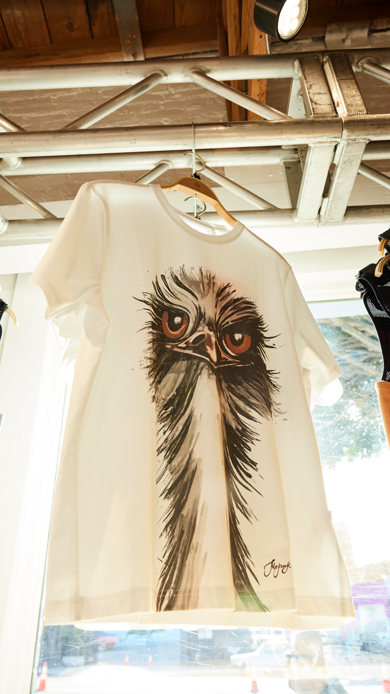 A white T-shirt hanging on a wooden hanger, featuring a large, artistic illustration of an ostrichs face with expressive eyes. Sunlight streams in from a window behind the shirt.