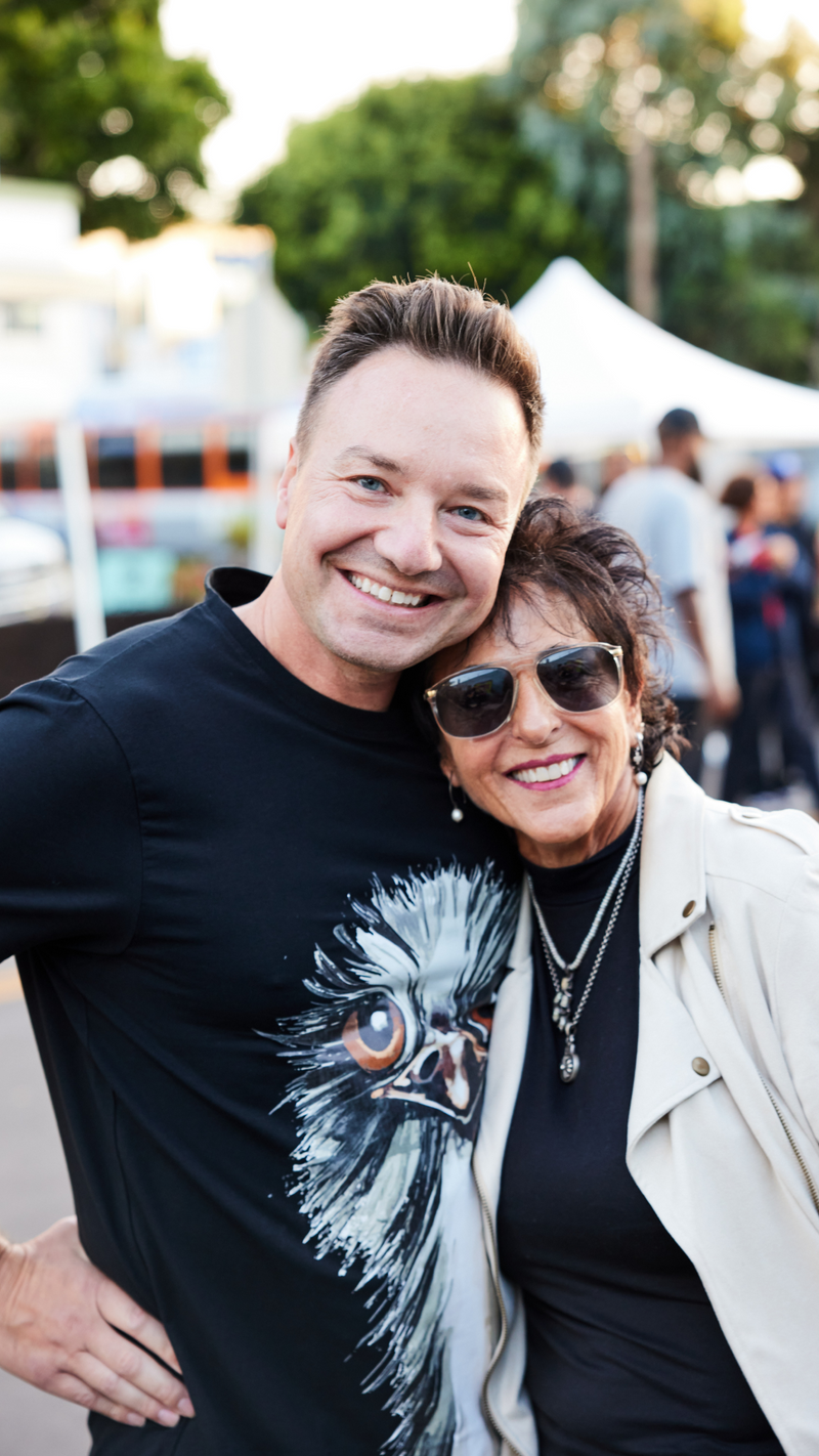 Two people smiling and posing together outdoors at an event. The man wears a black shirt with an ostrich face, and the woman wears sunglasses and a light jacket. There are blurred people and tents in the background.