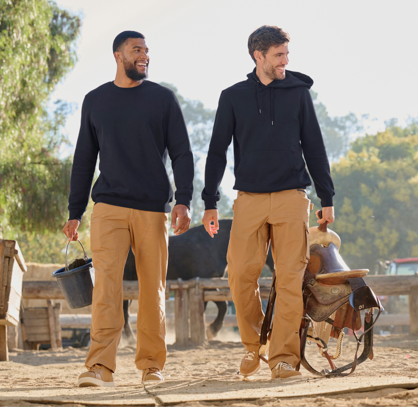 Two men wearing black sweatshirts and tan pants walk outdoors on a ranch. One carries a bucket, and the other holds a saddle. They are smiling, surrounded by fences, trees, and a blurred horse in the background.