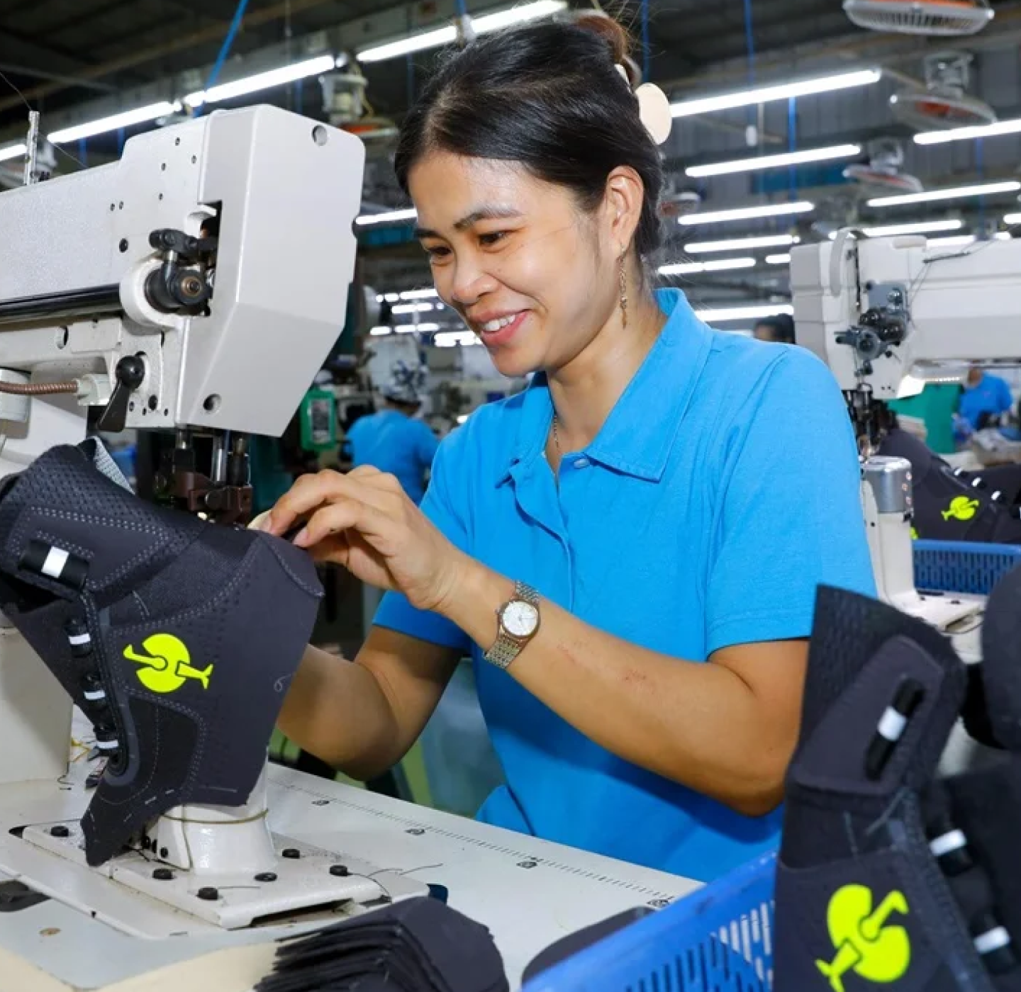 A woman in a blue polo shirt smiles while sewing black fabric pieces with green logos at a workstation in a busy garment factory.