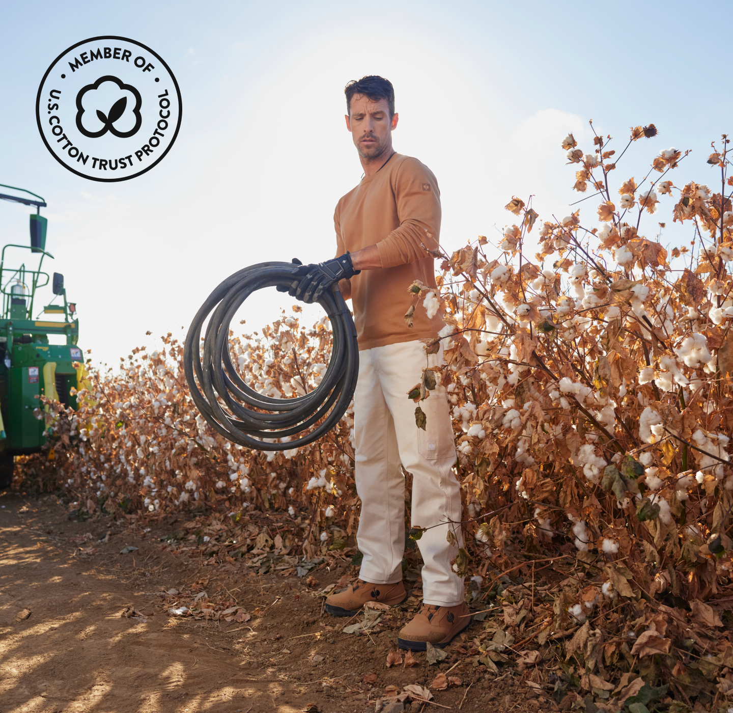 A man in work clothes and gloves holds a black hose in a cotton field near a green tractor. There is a Member of U.S. Cotton Trust Protocol logo in the top left corner.