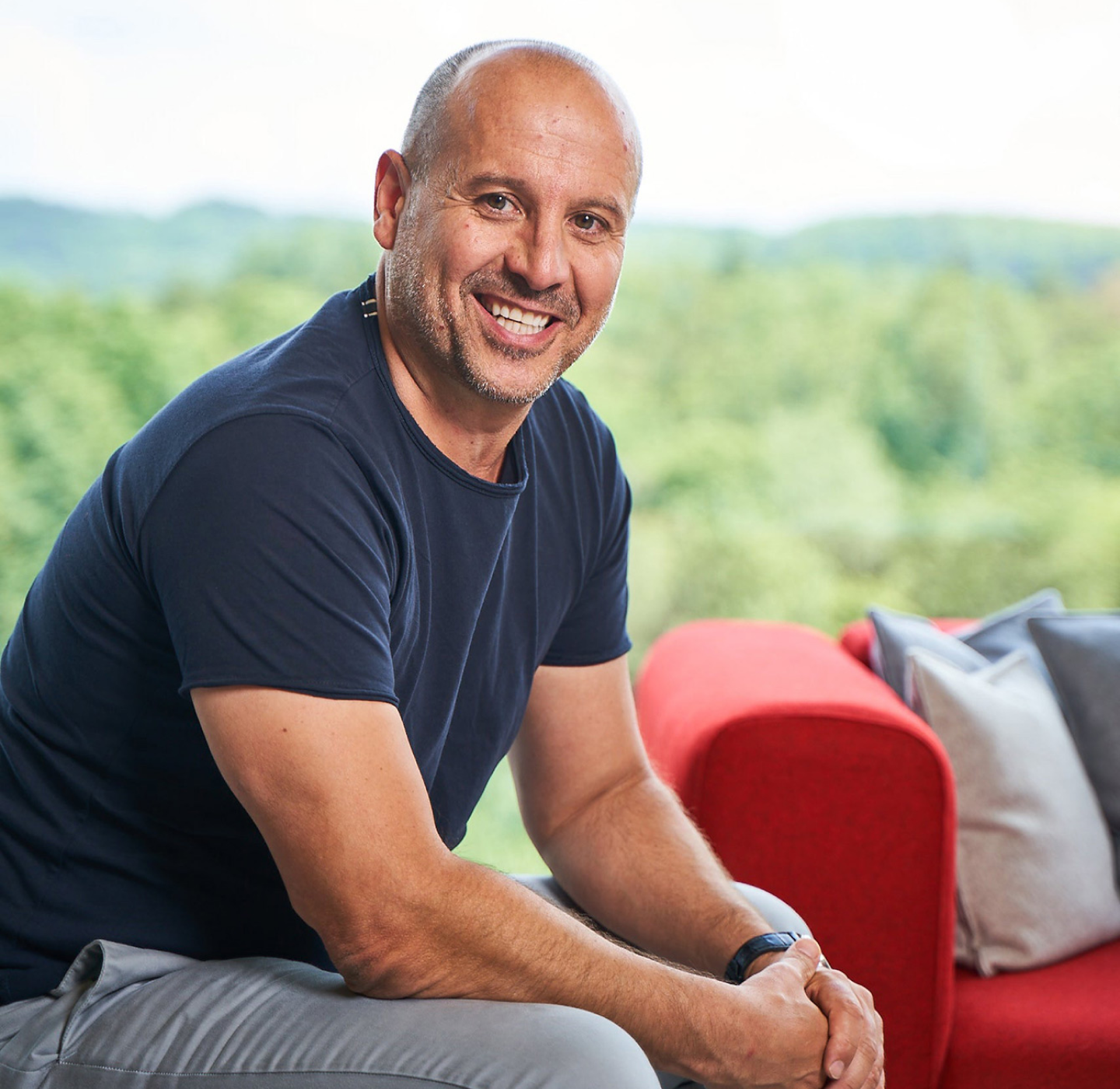 A smiling man with a shaved head, wearing a dark t-shirt and light pants, sits on a red sofa with pillows. Green trees and a bright sky are visible through a large window behind him.