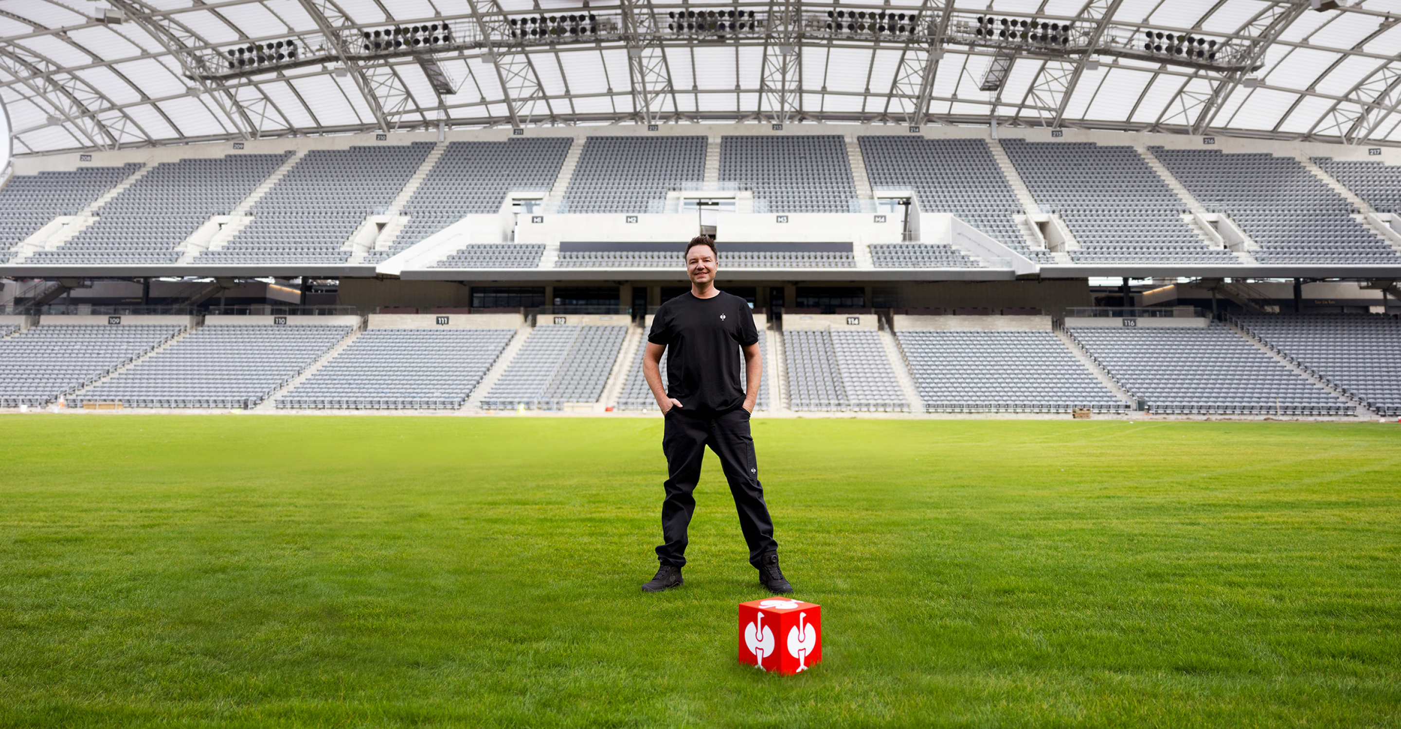 A person in black clothes stands on a soccer field, smiling, with a red block showing white chicken drumstick icons at their feet. The stadium seating and roof are visible in the background.