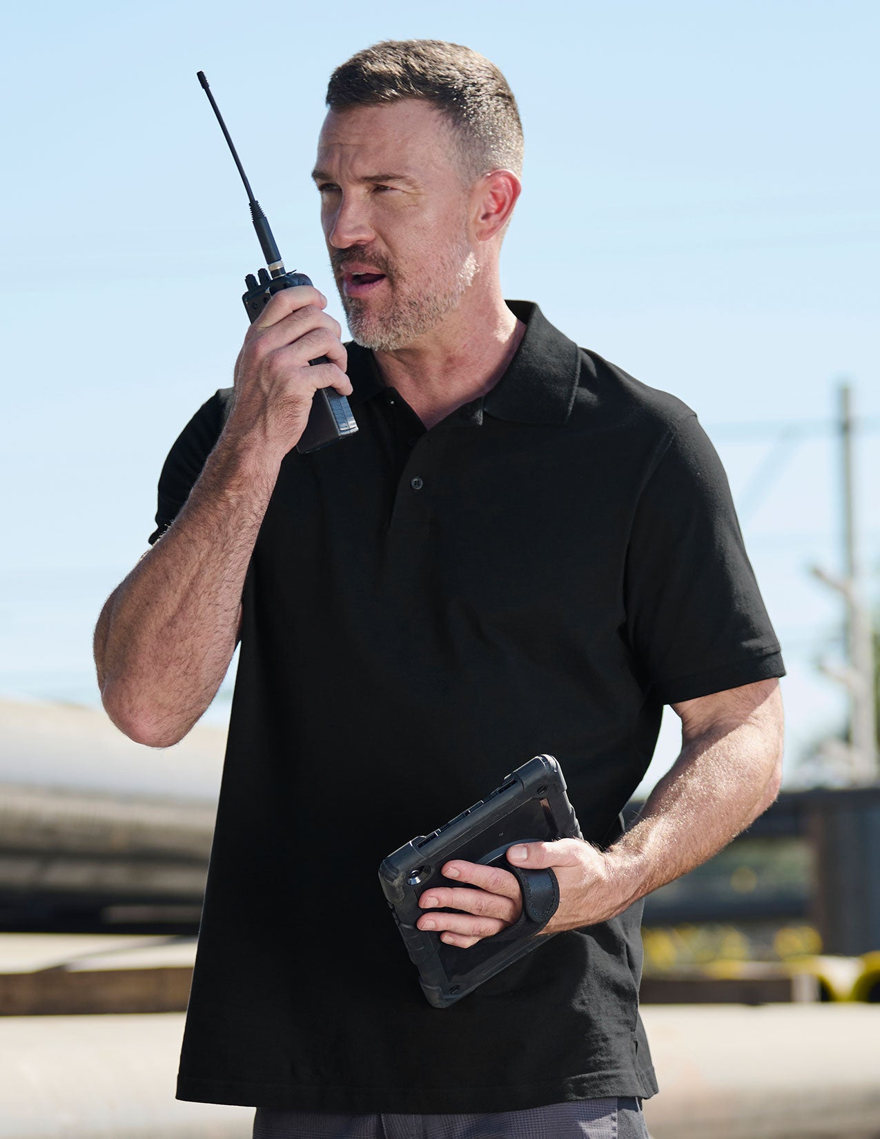 Man with a black polo shirt holding a walkie-talkie and a tablet outdoors.