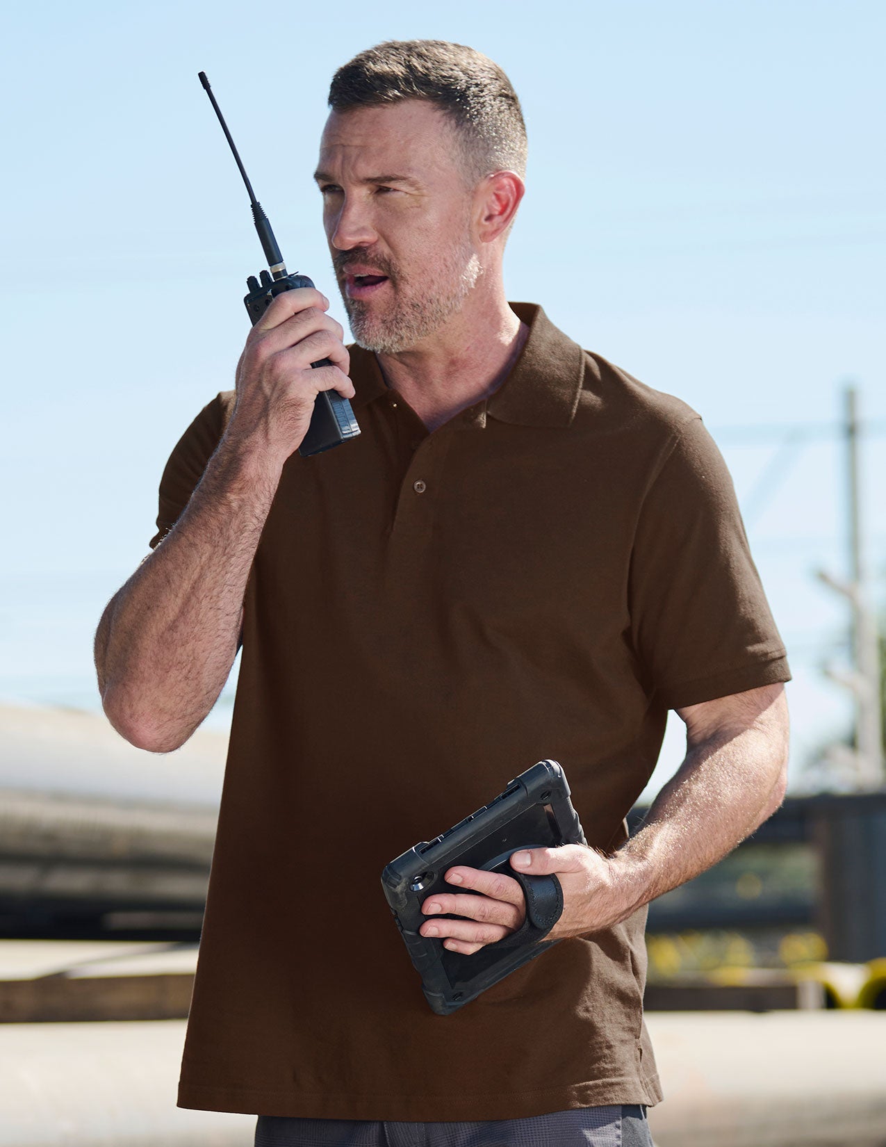 Man in brown polo shirt holding a walkie-talkie and tablet outdoors.
