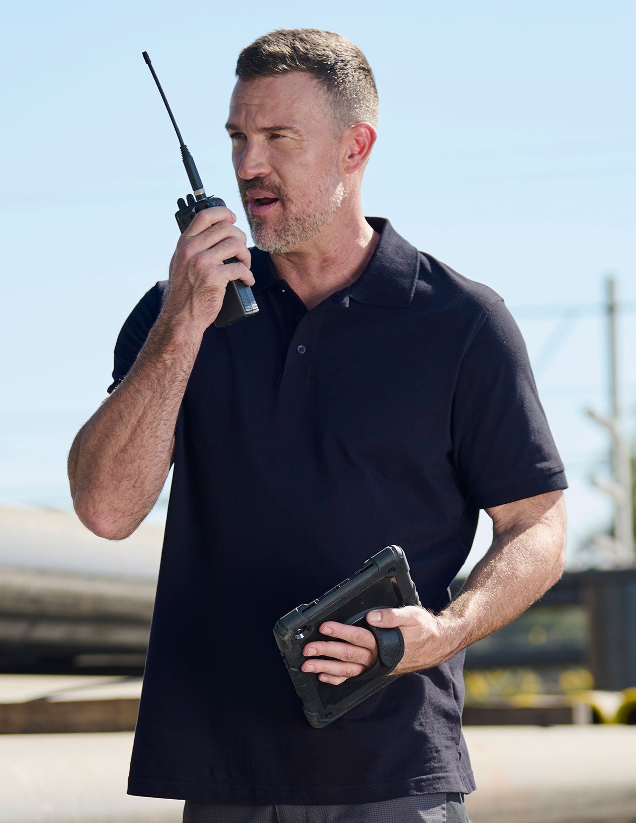 Man in neroblue shirt holding a walkie-talkie and tablet outdoors.