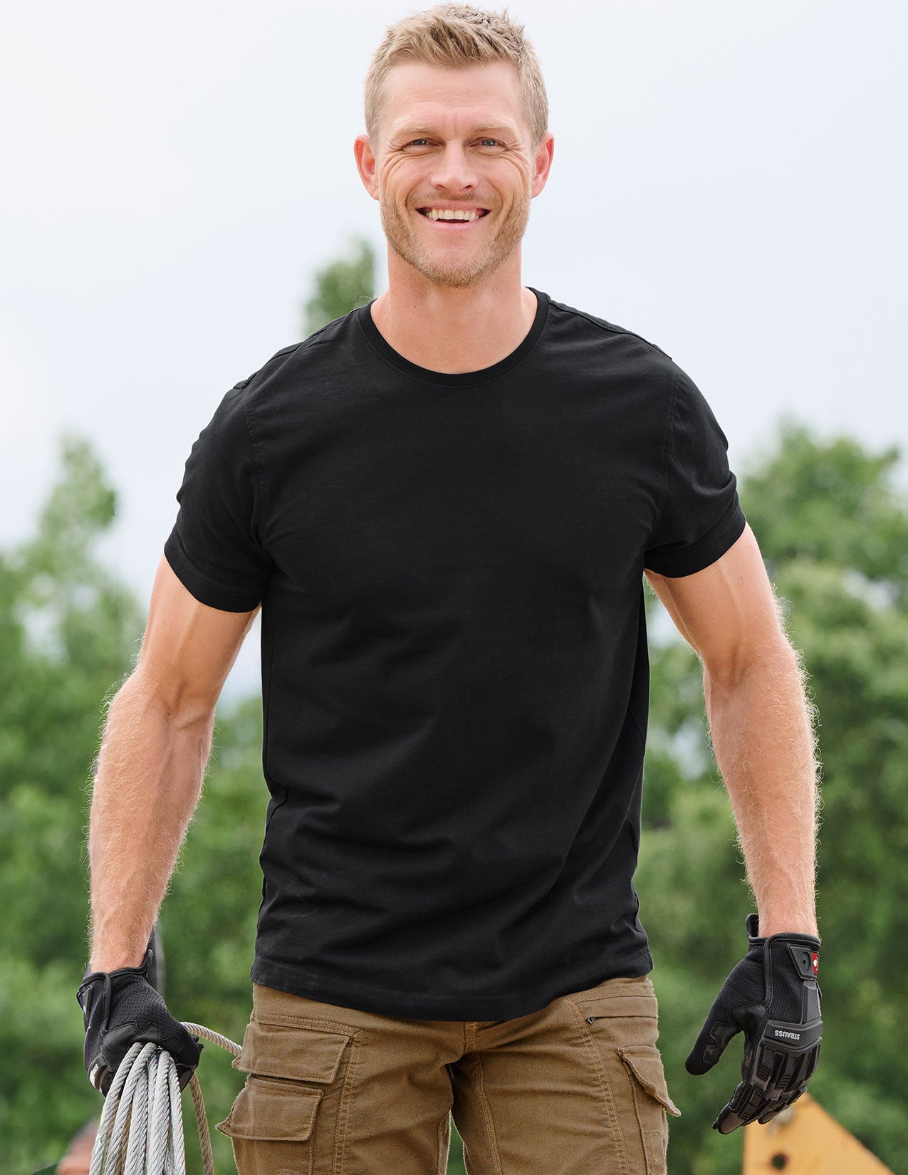 Man wearing a black t-shirt and gloves outdoors with greenery in the background.
