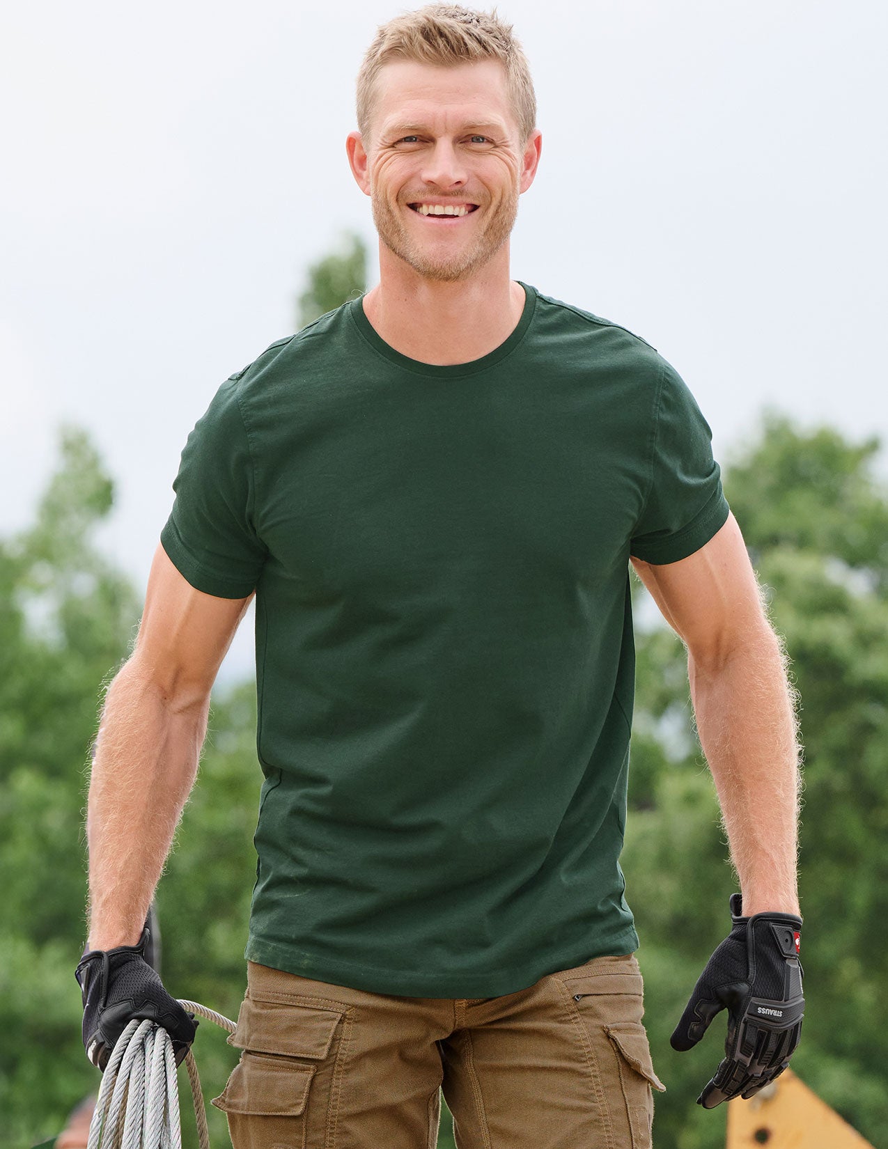 Man wearing a green t-shirt and black gloves outdoors with trees in the background.