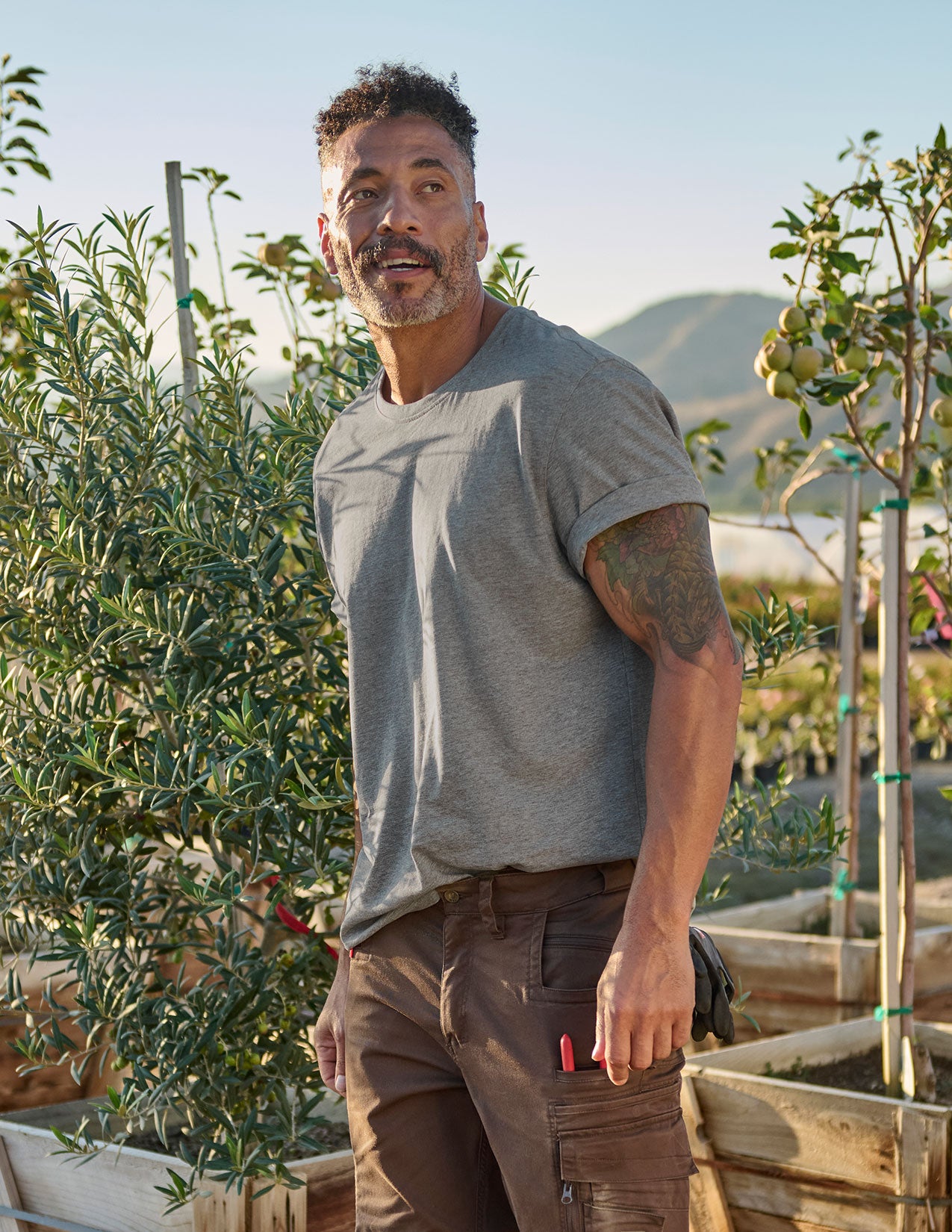 Man standing in a garden with plants and mountains in the background.