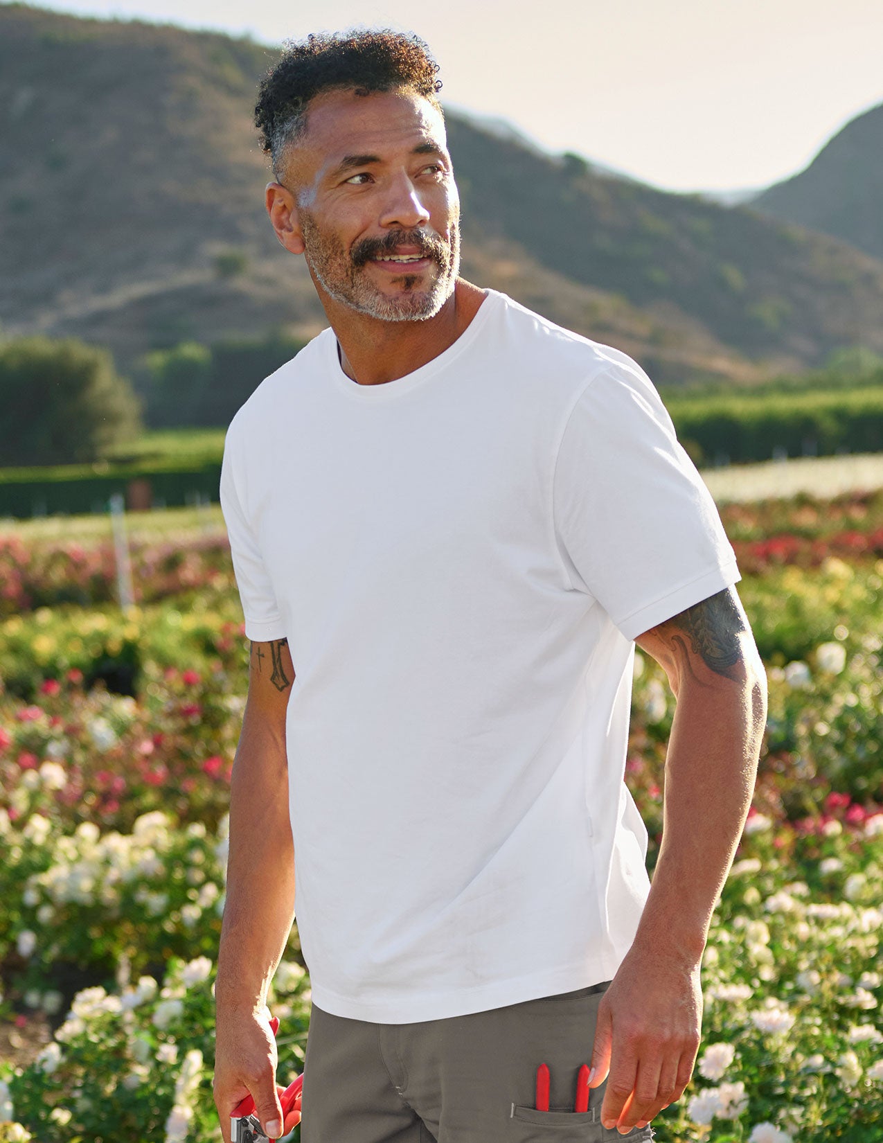 Man wearing a white t-shirt standing in a field with mountains in the background.