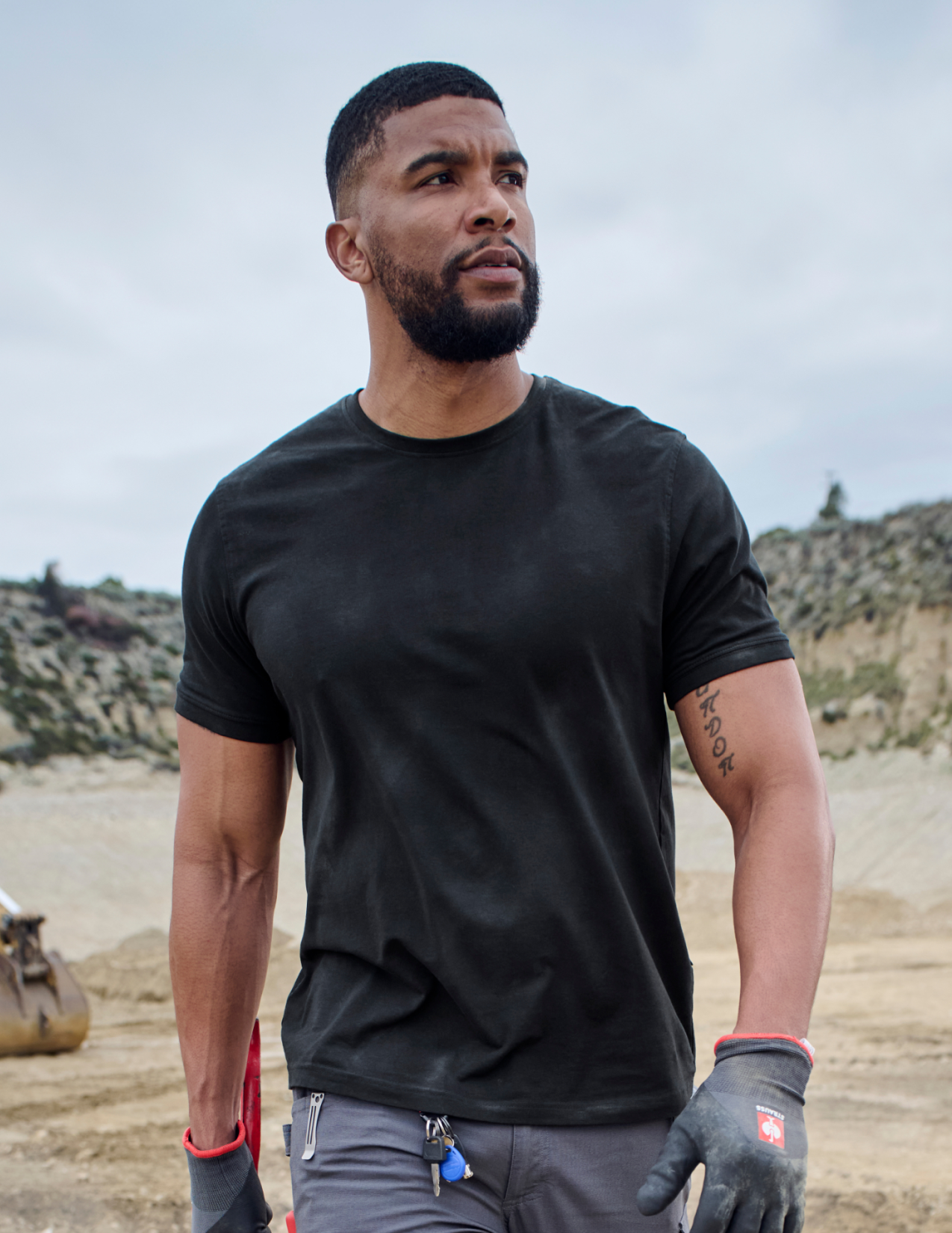 Man wearing a black t-shirt and gloves standing outdoors with a mountainous background