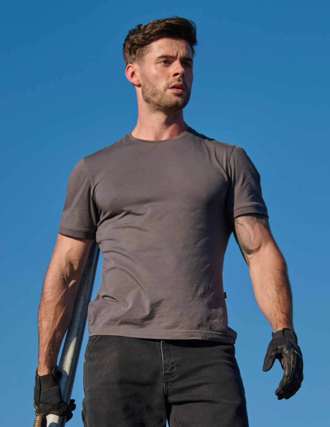 Man wearing a gray t-shirt and black gloves against a clear blue sky