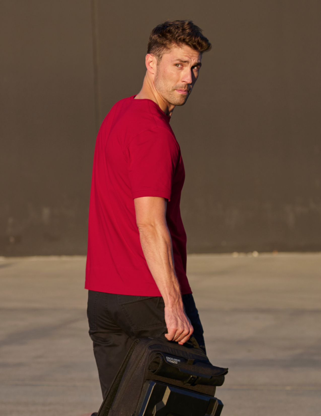 Man in a red shirt holding a black bag against a plain background