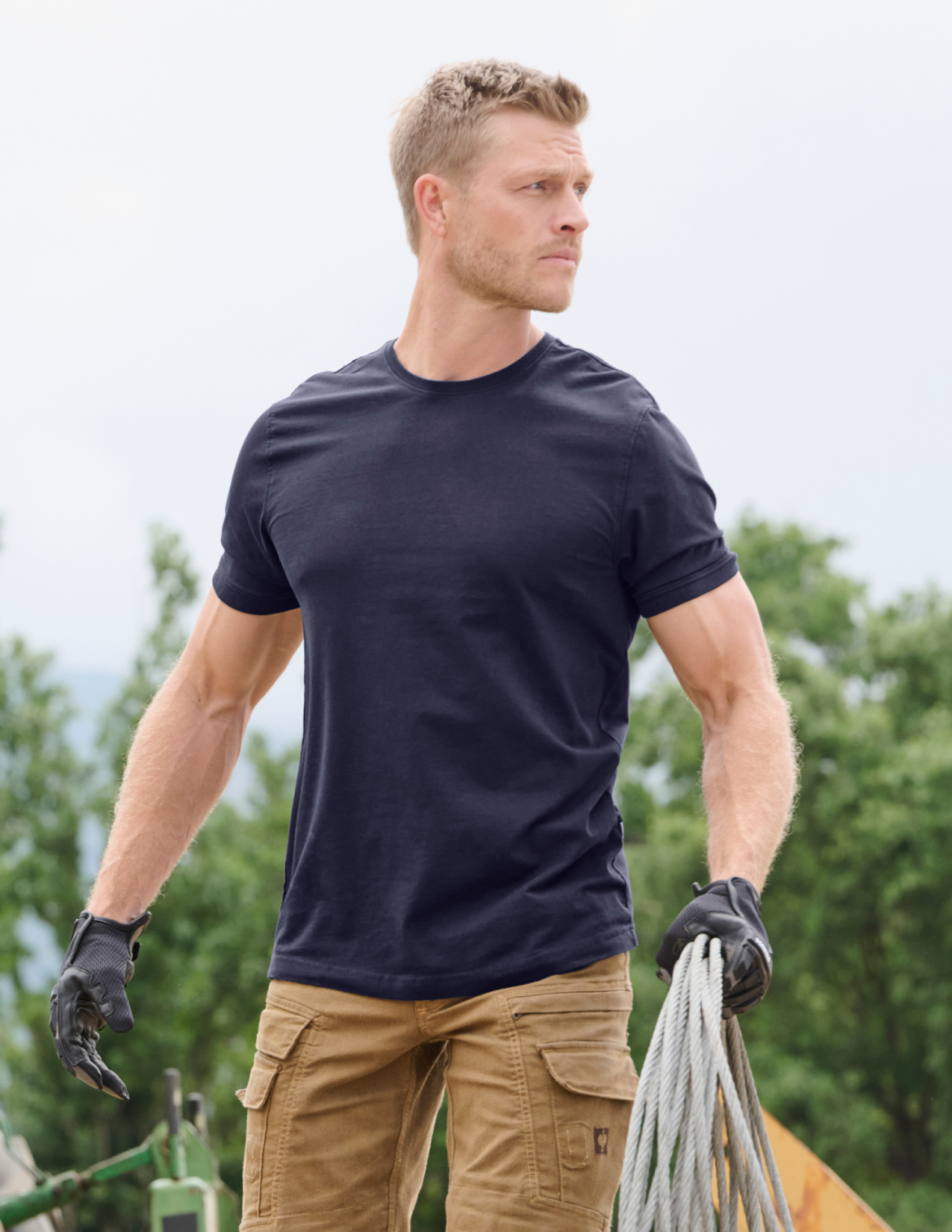 Man wearing a navy t-shirt and beige pants holding coiled cable outdoors.