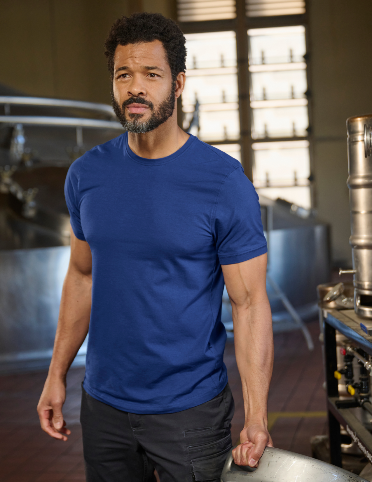 Man wearing a blue t-shirt in an brewery setting with equipment in the background