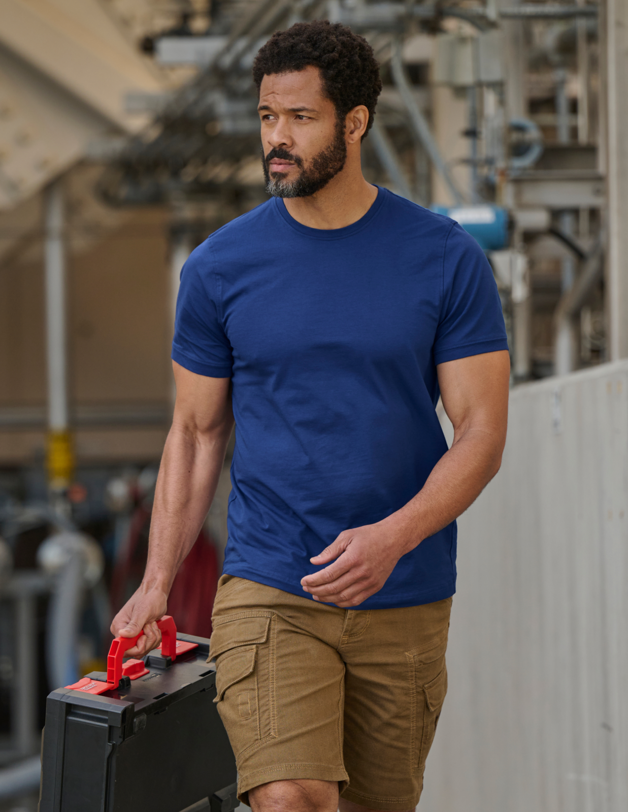 Man in a blue shirt and beige shorts carrying a toolbox in an industrial setting