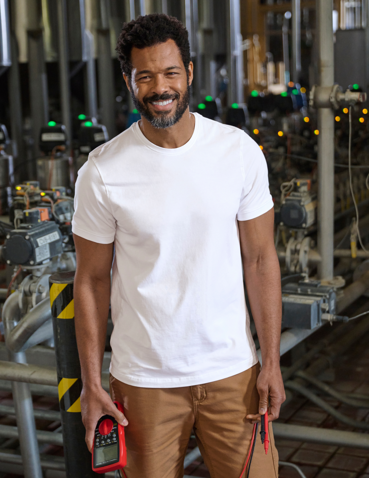 Man in a white t-shirt and brown pants holding a device in an industrial setting