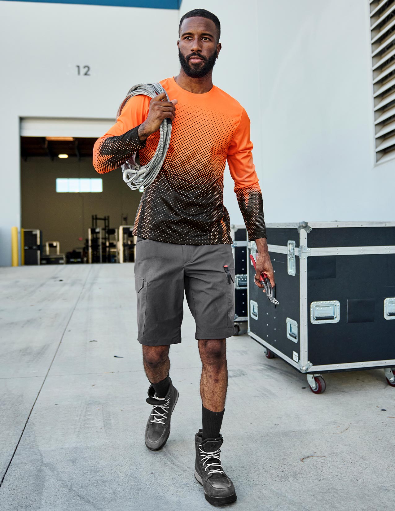 Man in orange shirt and gray shorts holding a tool in an industrial setting