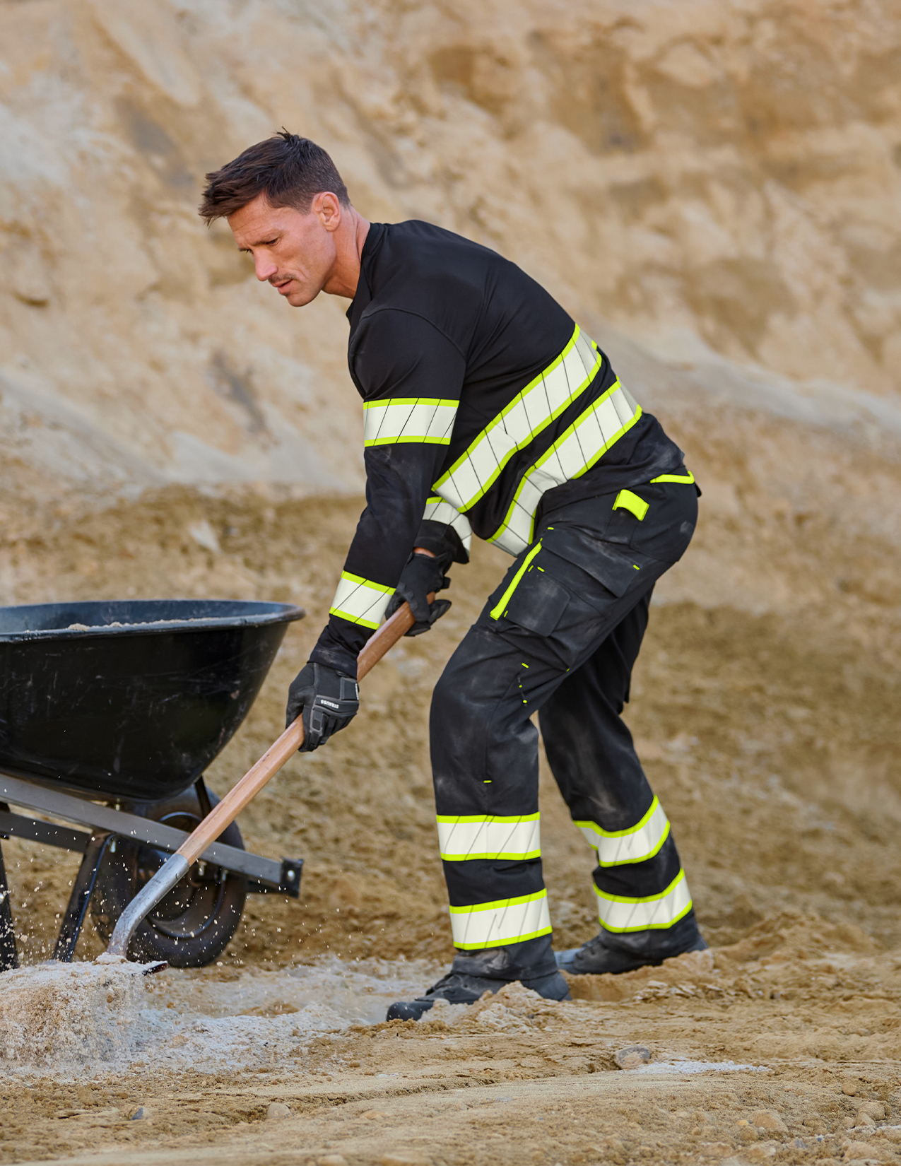 Person in high-visibility workwear shoveling sand with a wheelbarrow in the background