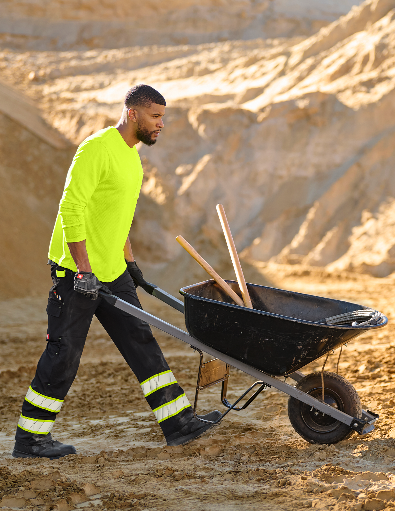 Person in high-visibility clothing pushing a wheelbarrow with tools on a construction site.