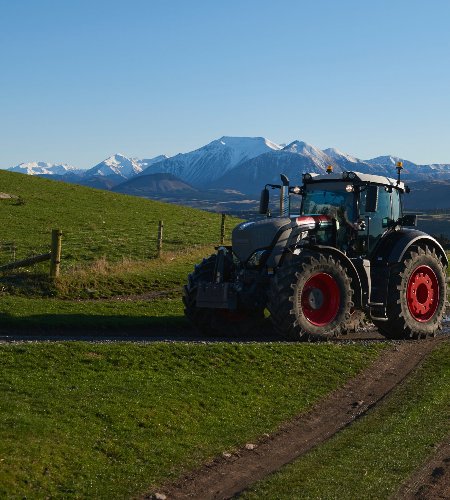 Tractor driving on a gravel path through green hills with mountains in the background.