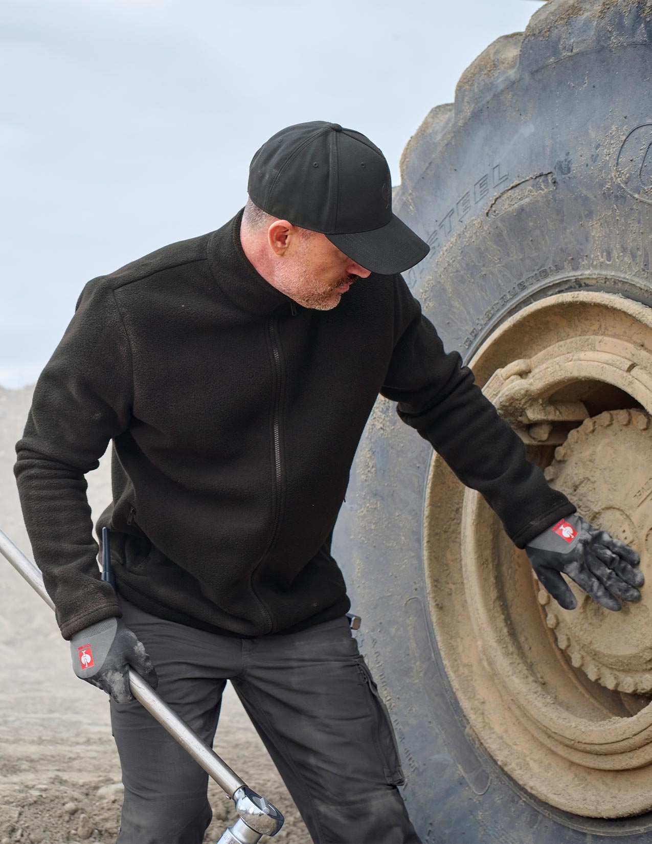 Person in black jacket and cap inspecting a large tire on a vehicle.